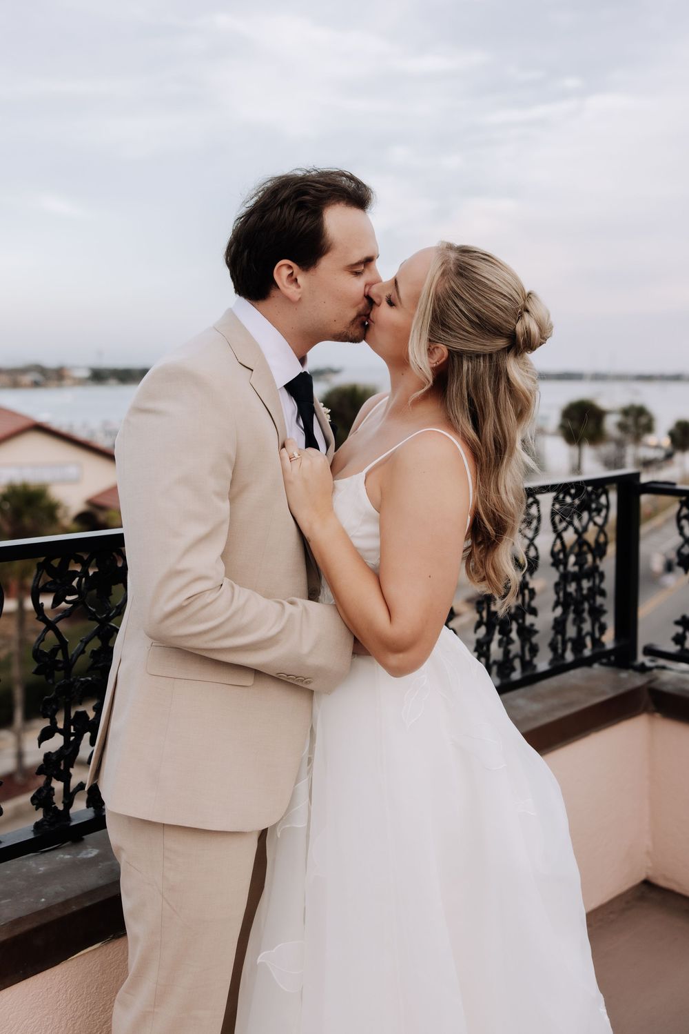 A bride and groom share a romantic kiss on a balcony overlooking the water, she in white gown, he in tan suit.