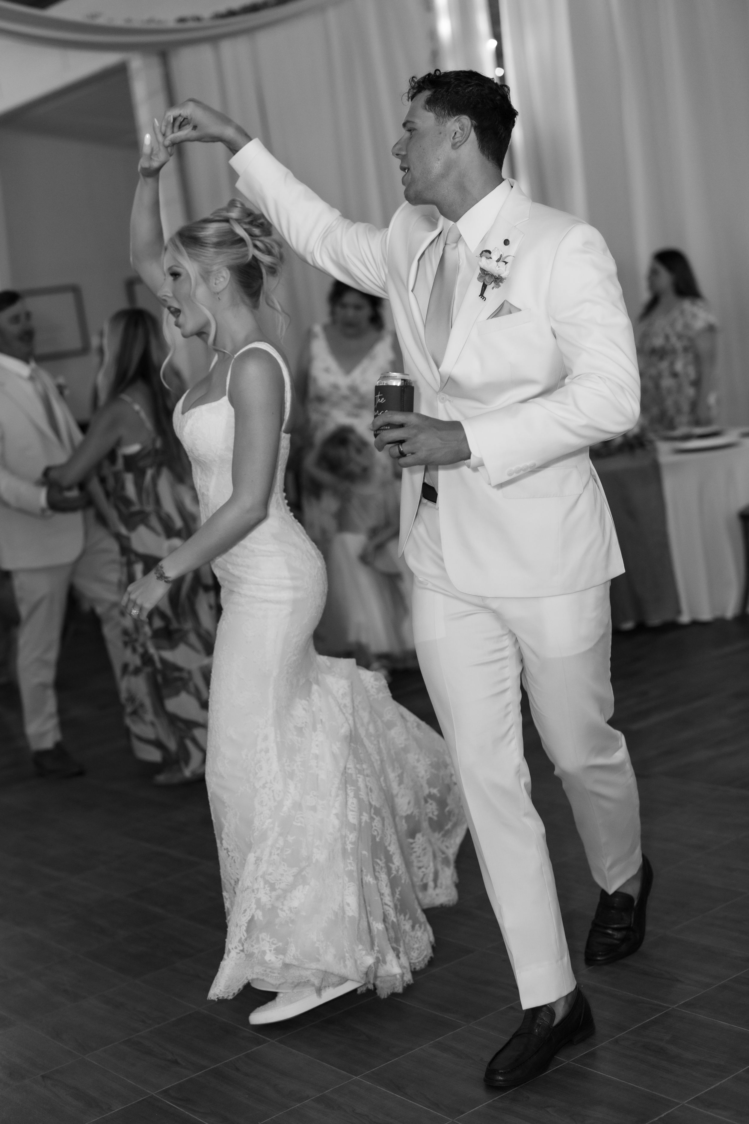 Black and white photo of bride and groom sharing first dance at wedding reception, groom in white suit twirling bride.