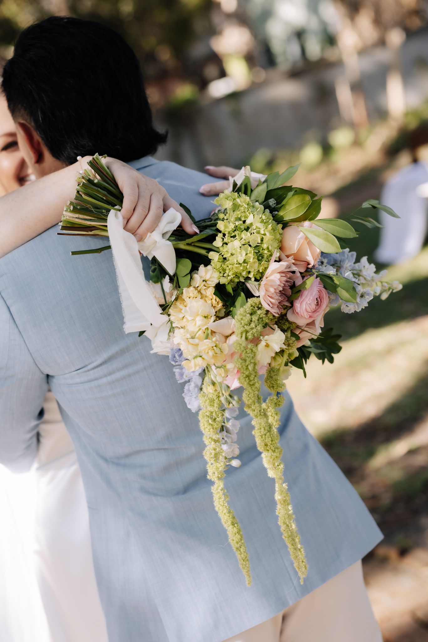 Bride holding a cascading wedding bouquet with pink roses and yellow flowers while embracing groom in light blue suit.