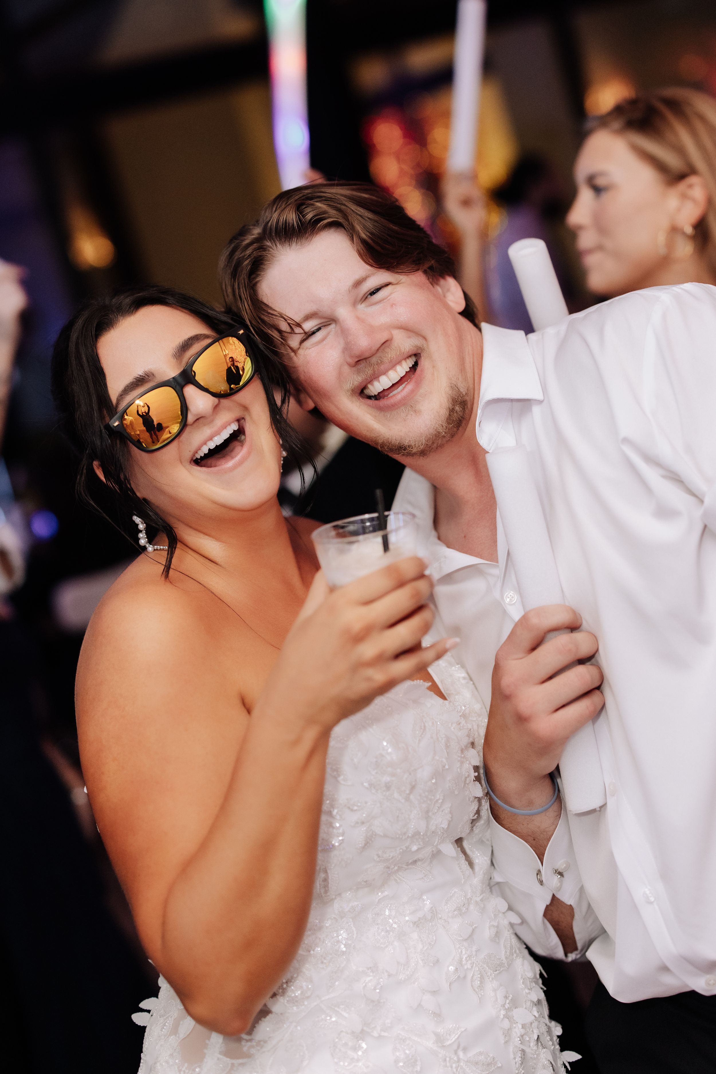 Laughing bride in sunglasses and groom in white shirt celebrate at wedding reception with champagne.