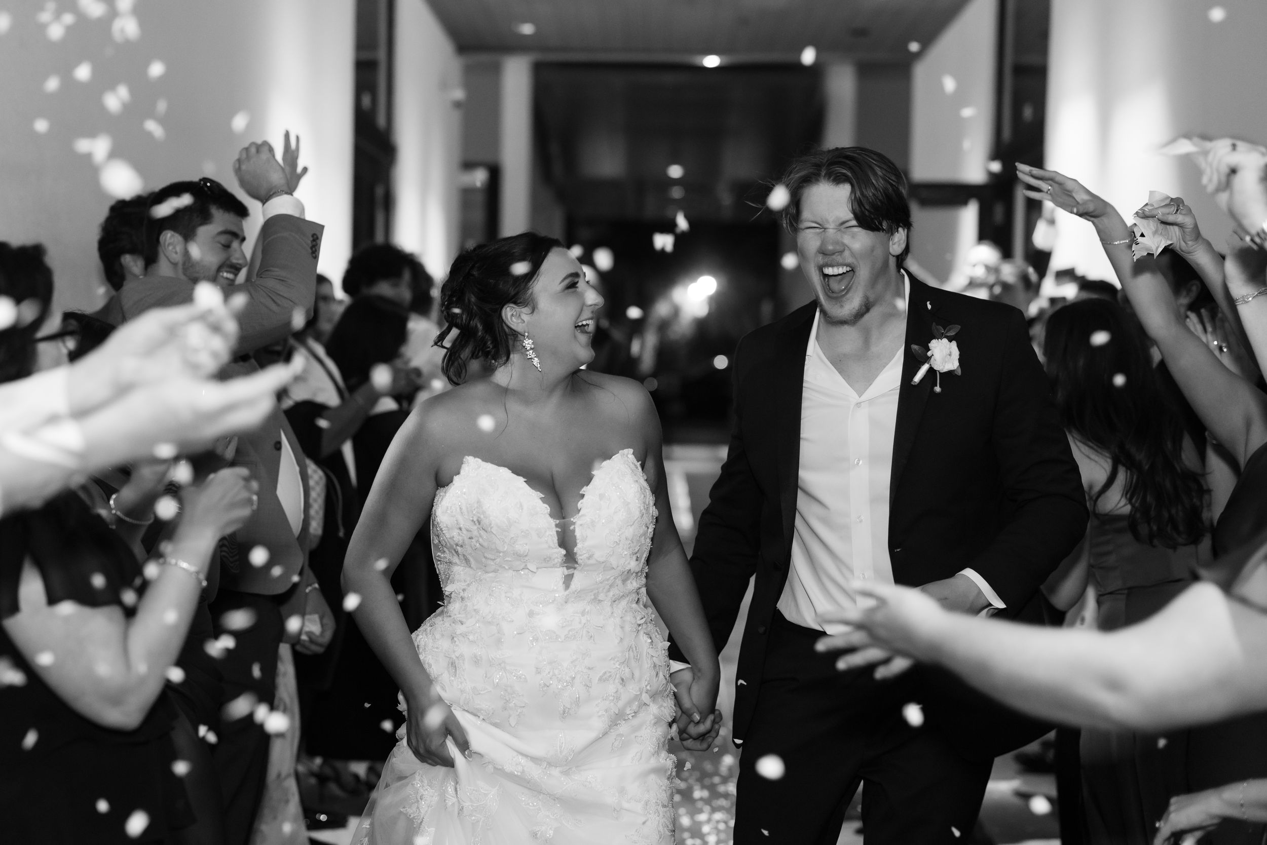 Black and white photo of a joyful bride and groom walking through a confetti shower surrounded by cheering wedding guests.