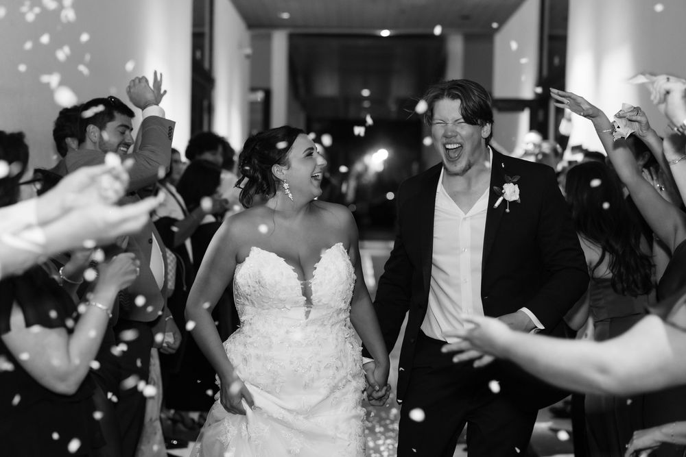 Black and white photo of a joyful bride and groom walking through a confetti shower surrounded by cheering wedding guests.