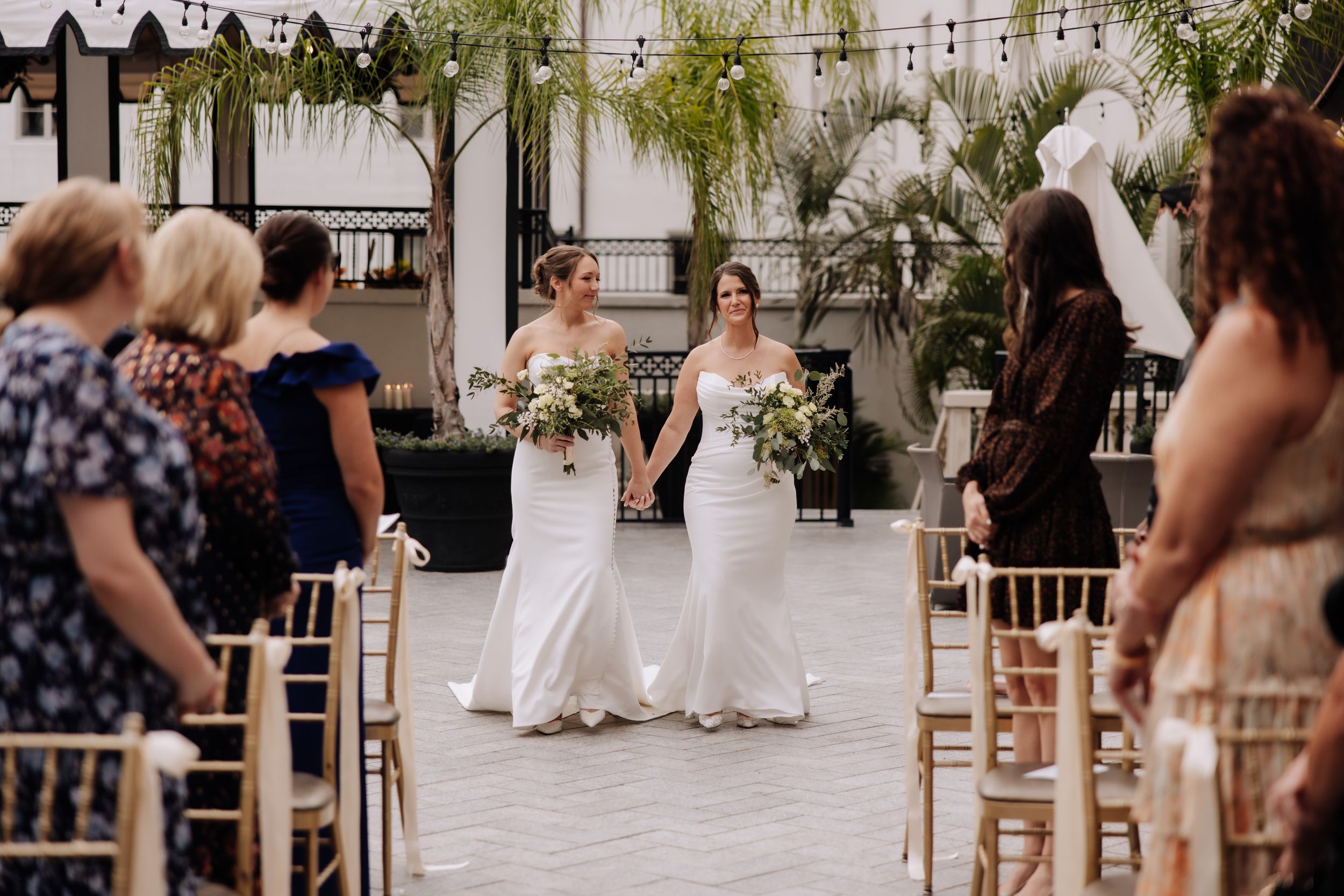 Two brides in white gowns hold bouquets and walk down the aisle at an elegant indoor wedding ceremony.