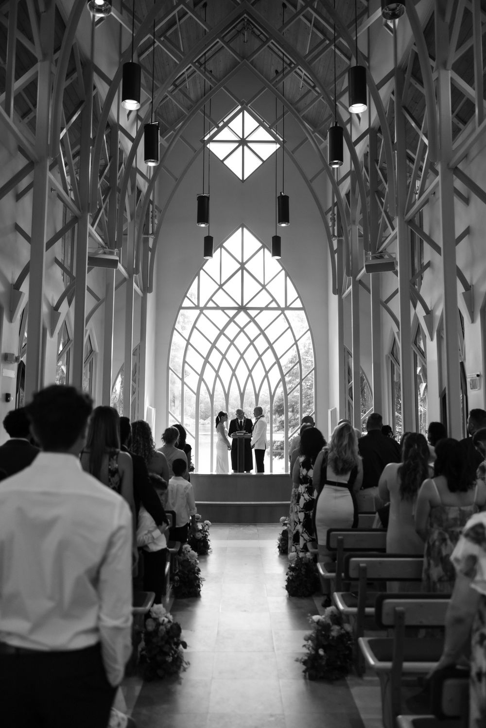Black and white photo of a wedding ceremony in a modern chapel with Gothic arches and large stained glass window.