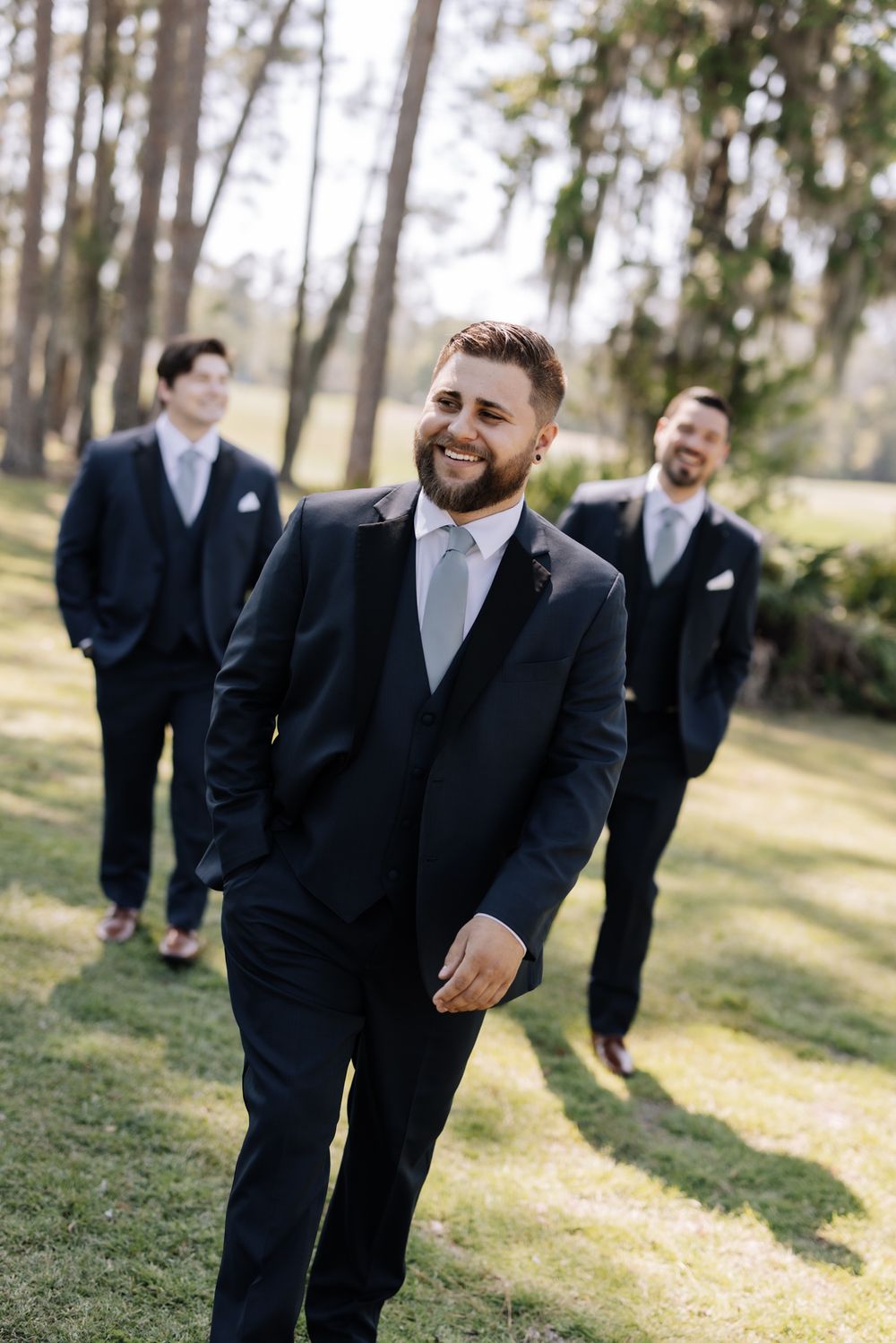 Groom smiling in navy suit with groomsmen in matching suits standing outdoors on green grass with trees in background.