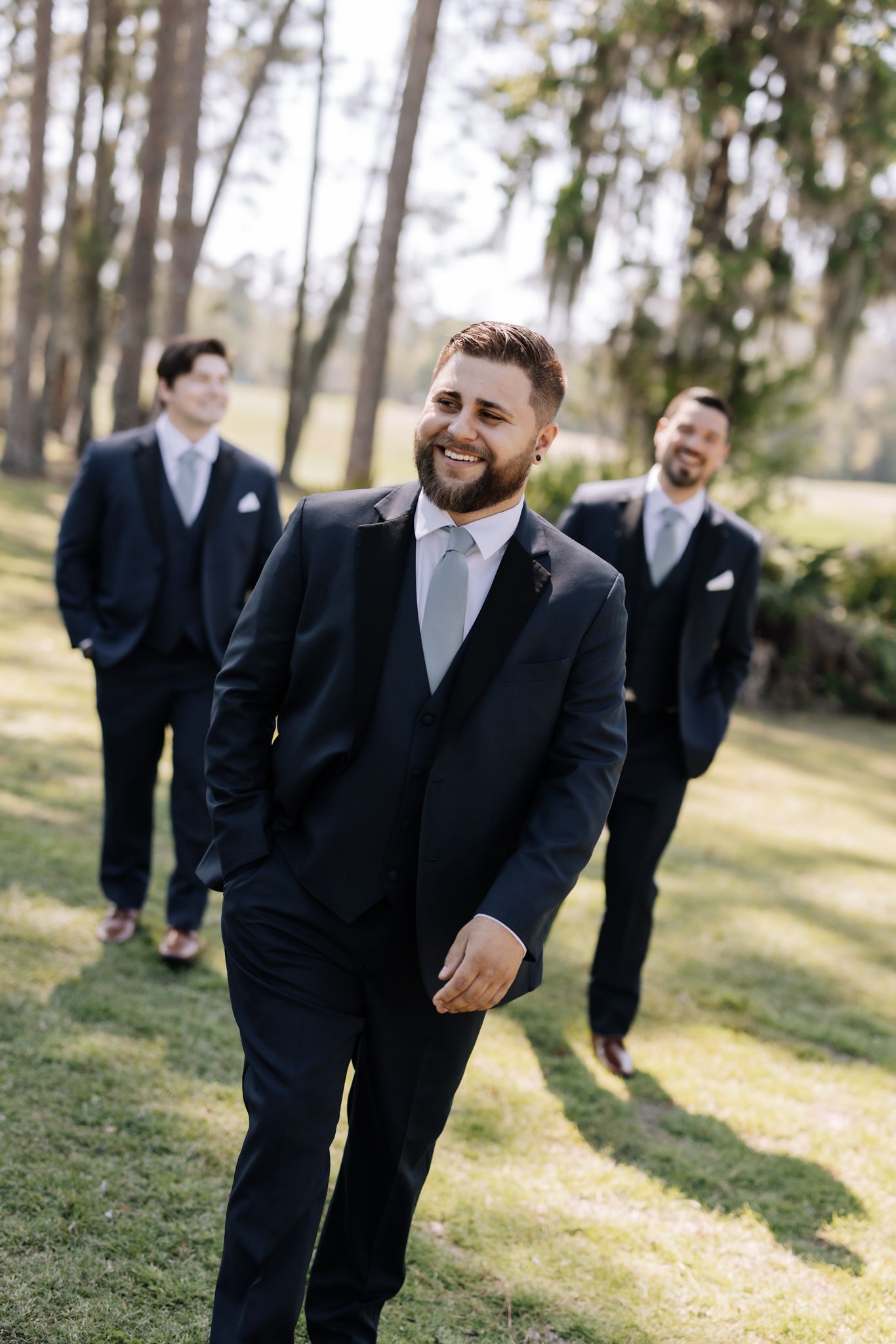 Groom smiling in navy suit with groomsmen in matching suits standing outdoors on green grass with trees in background.