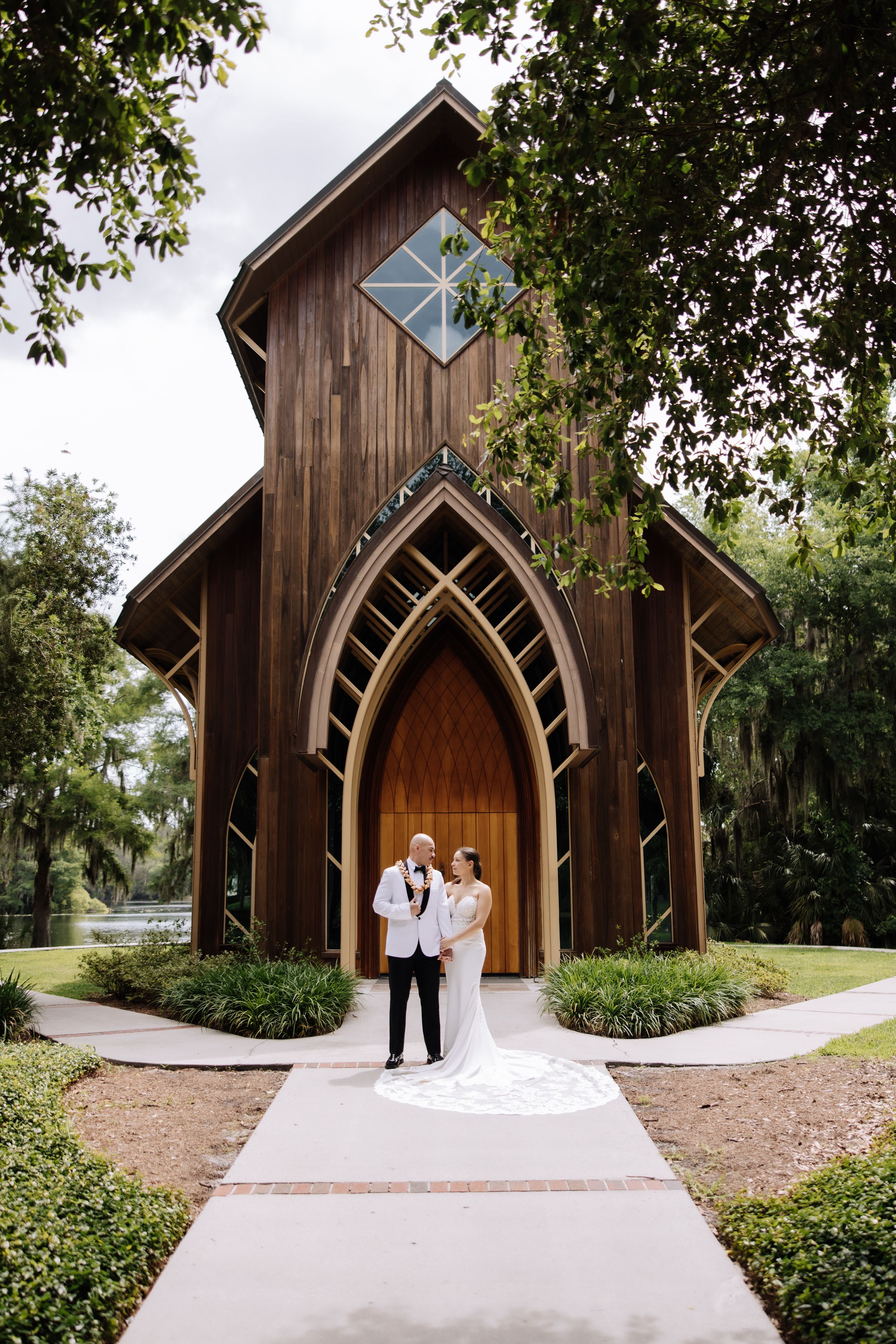 Bride and groom pose before a stunning rustic wooden chapel with Gothic arched doors on their wedding day.