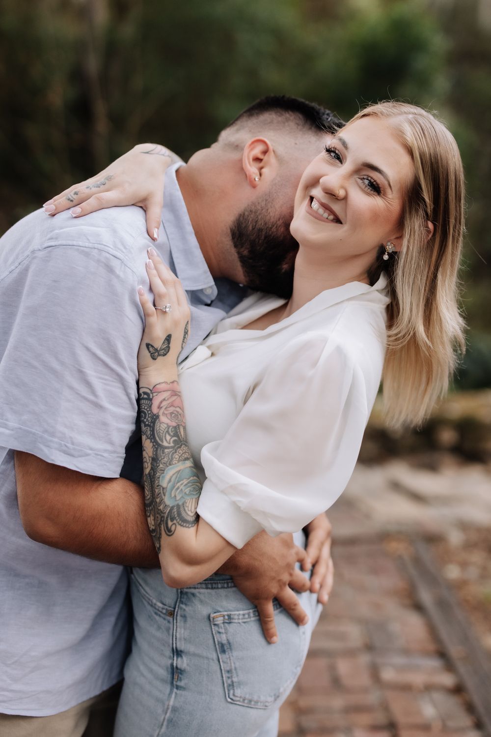A smiling blonde woman in a white blouse embraces a tattooed bearded man kissing her cheek outdoors.