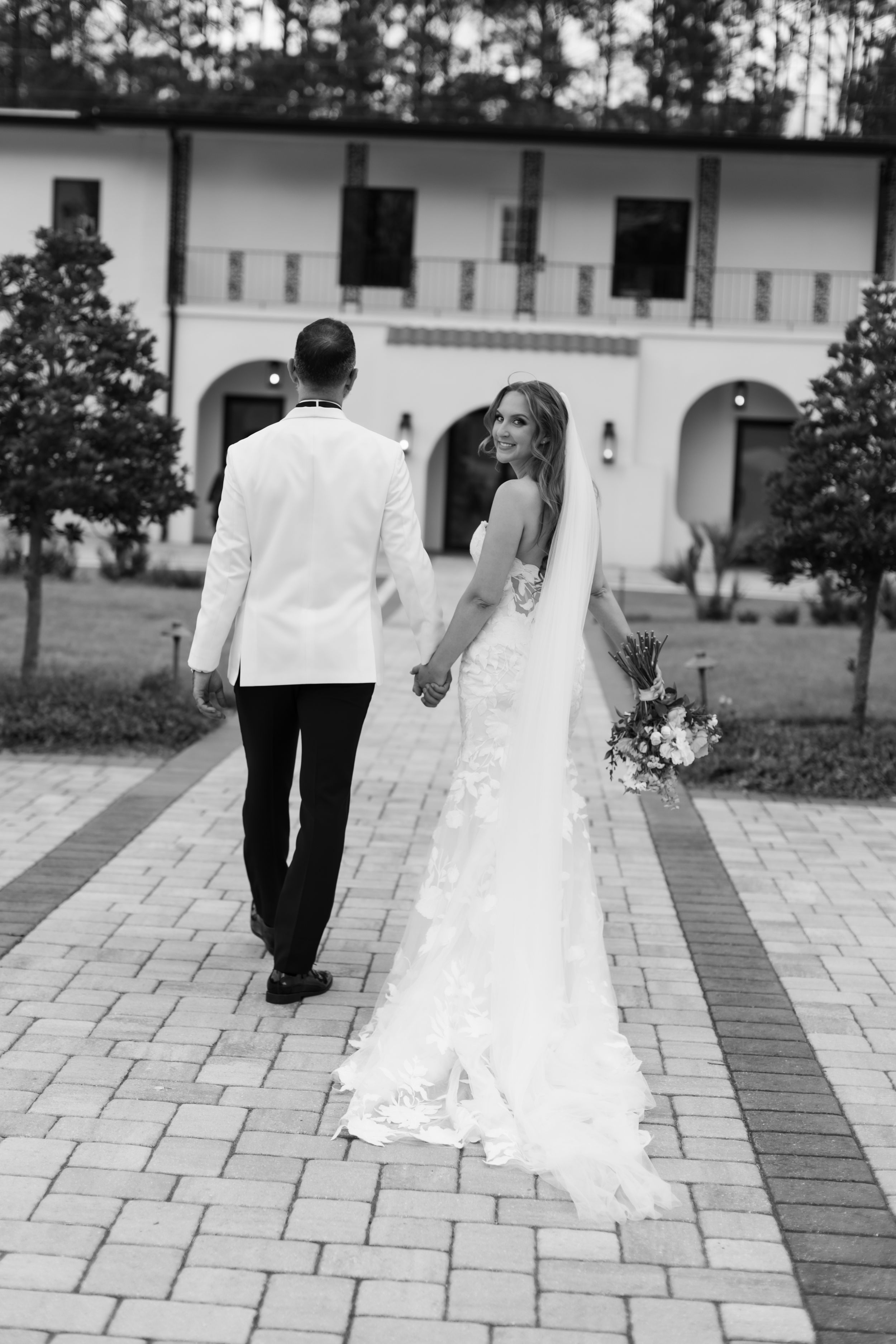 Black and white photo of bride and groom walking hand in hand toward elegant white venue, bride glancing back holding bouquet.