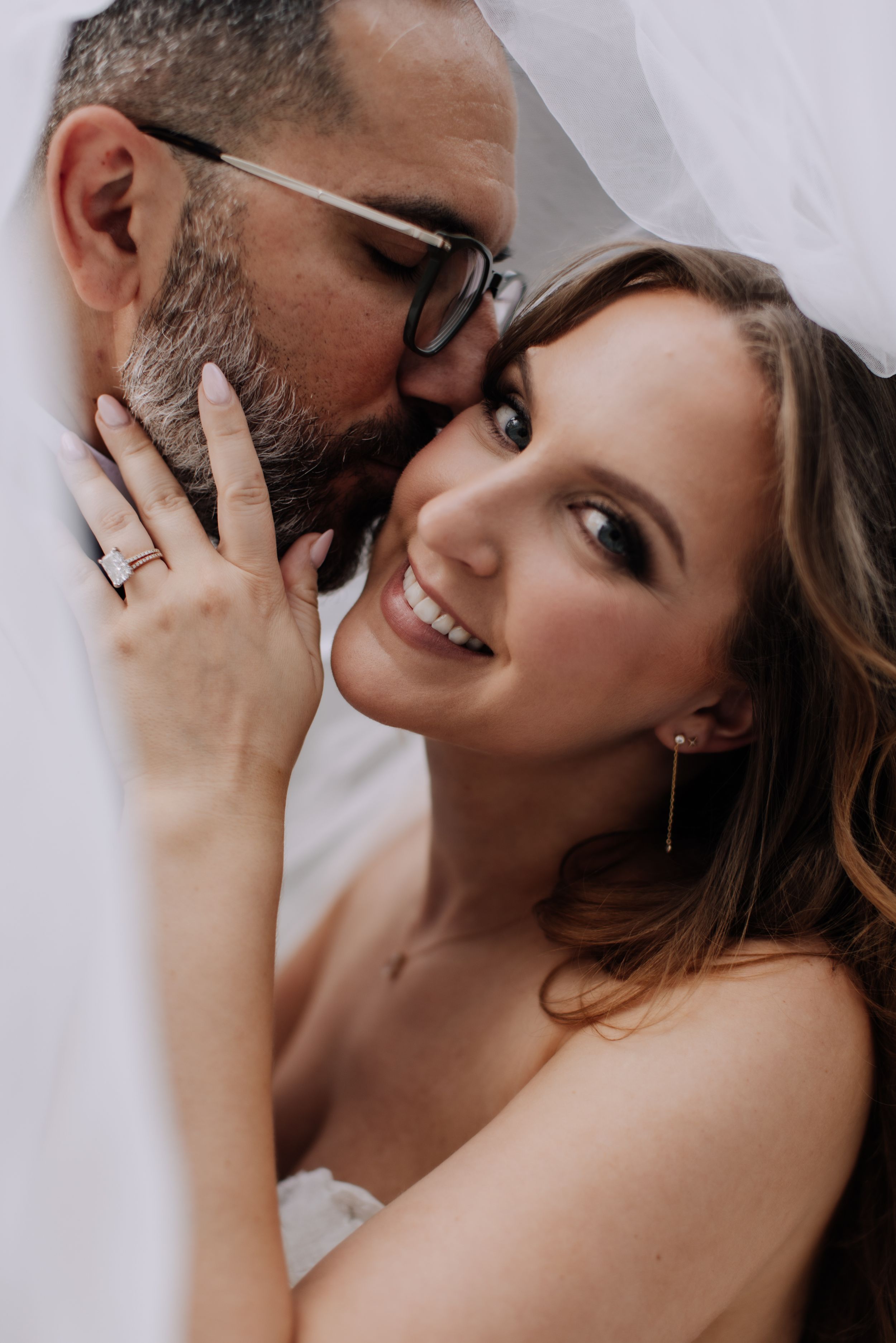 A smiling bride with a wedding ring poses as her bearded groom in glasses kisses her cheek under a white veil.