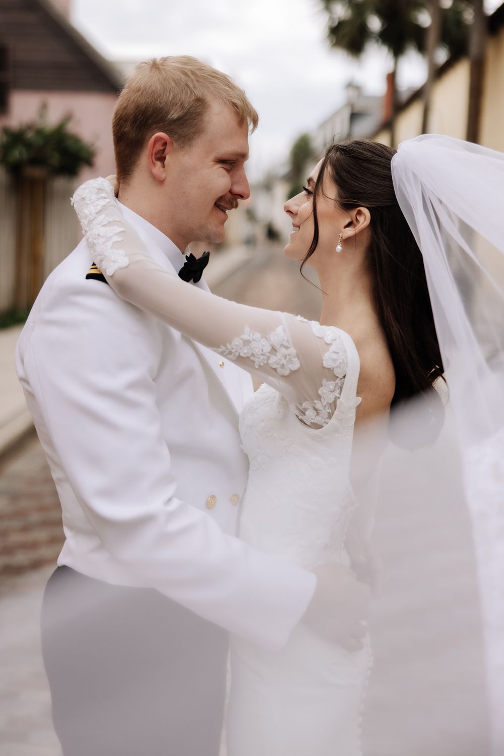 A smiling bride and groom in white wedding attire share a romantic moment on a cobblestone street.
