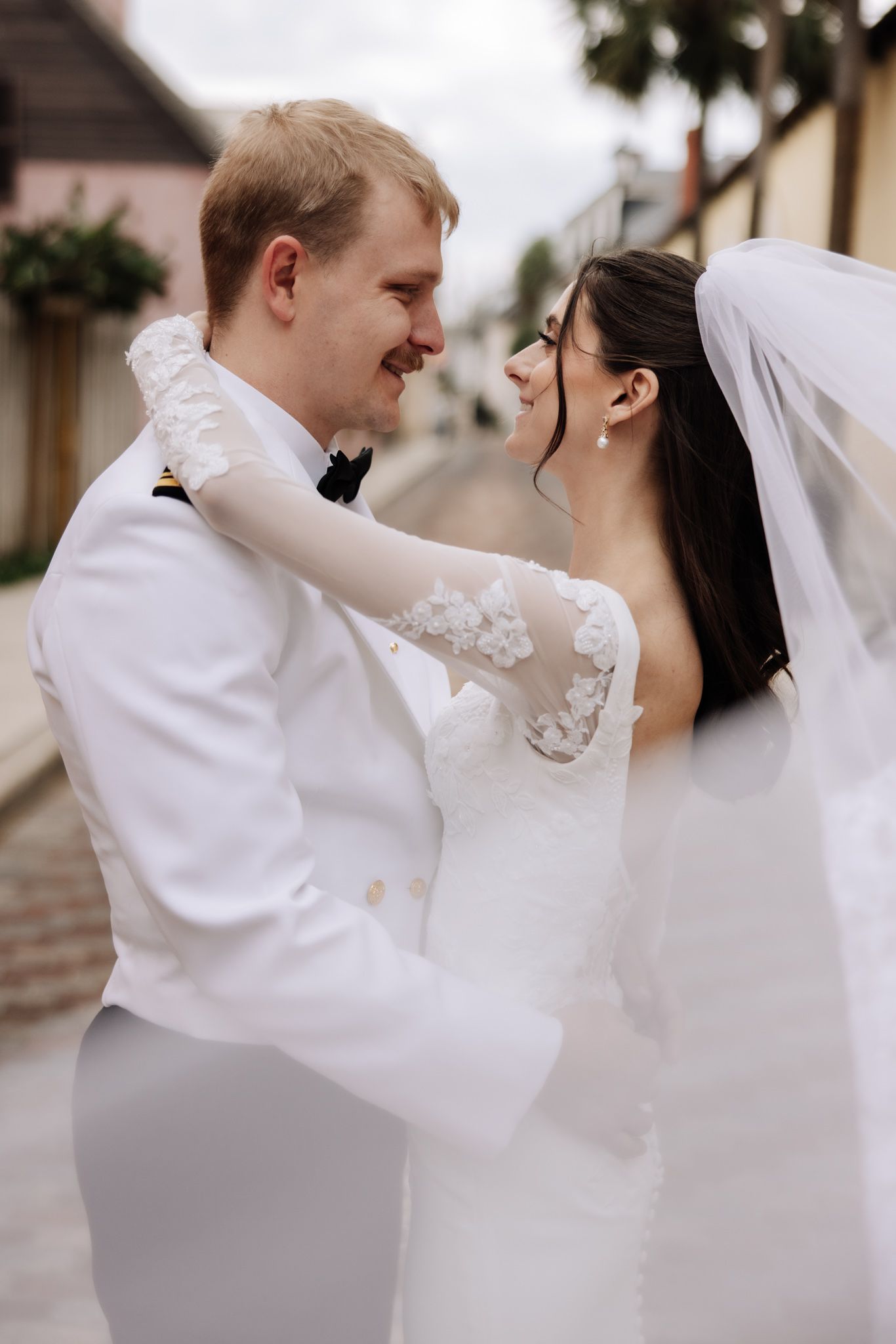 A smiling bride and groom in white wedding attire share a romantic moment on a cobblestone street.