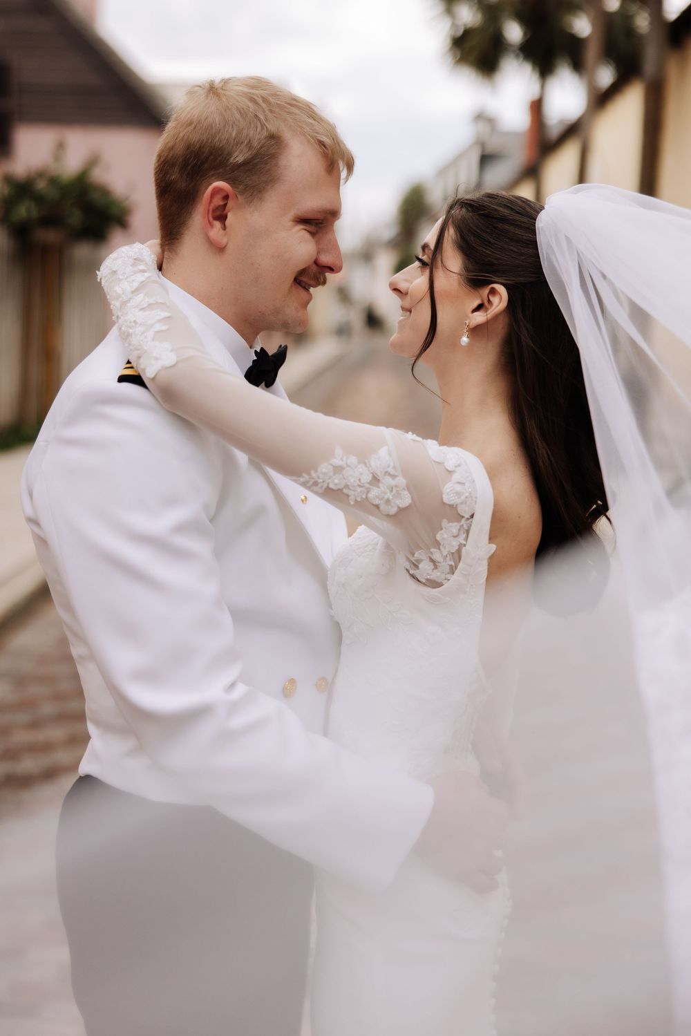 A smiling bride and groom in white wedding attire share a romantic moment on a cobblestone street.