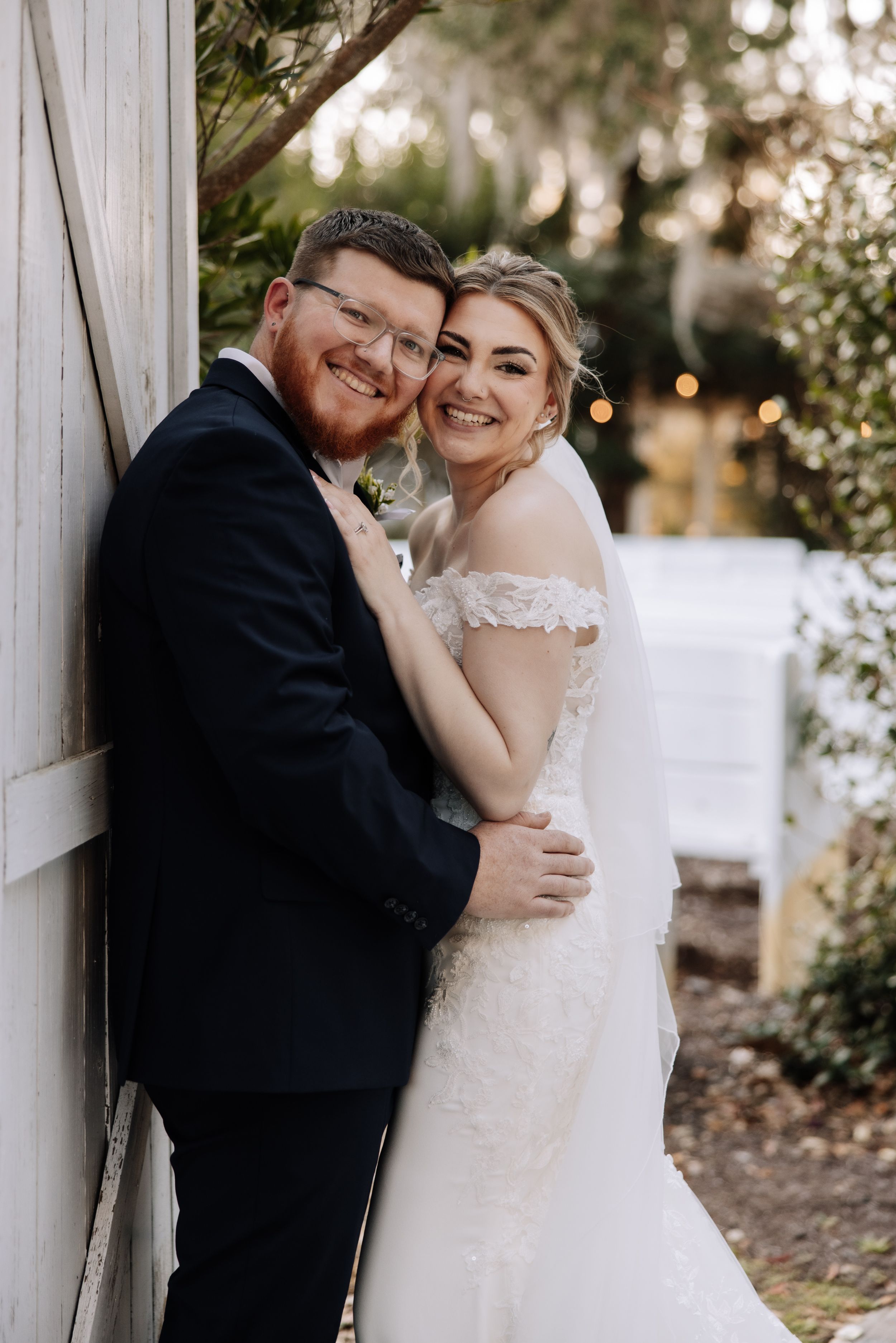 A smiling bride and groom in wedding attire embrace outdoors against a white barn wall surrounded by greenery.
