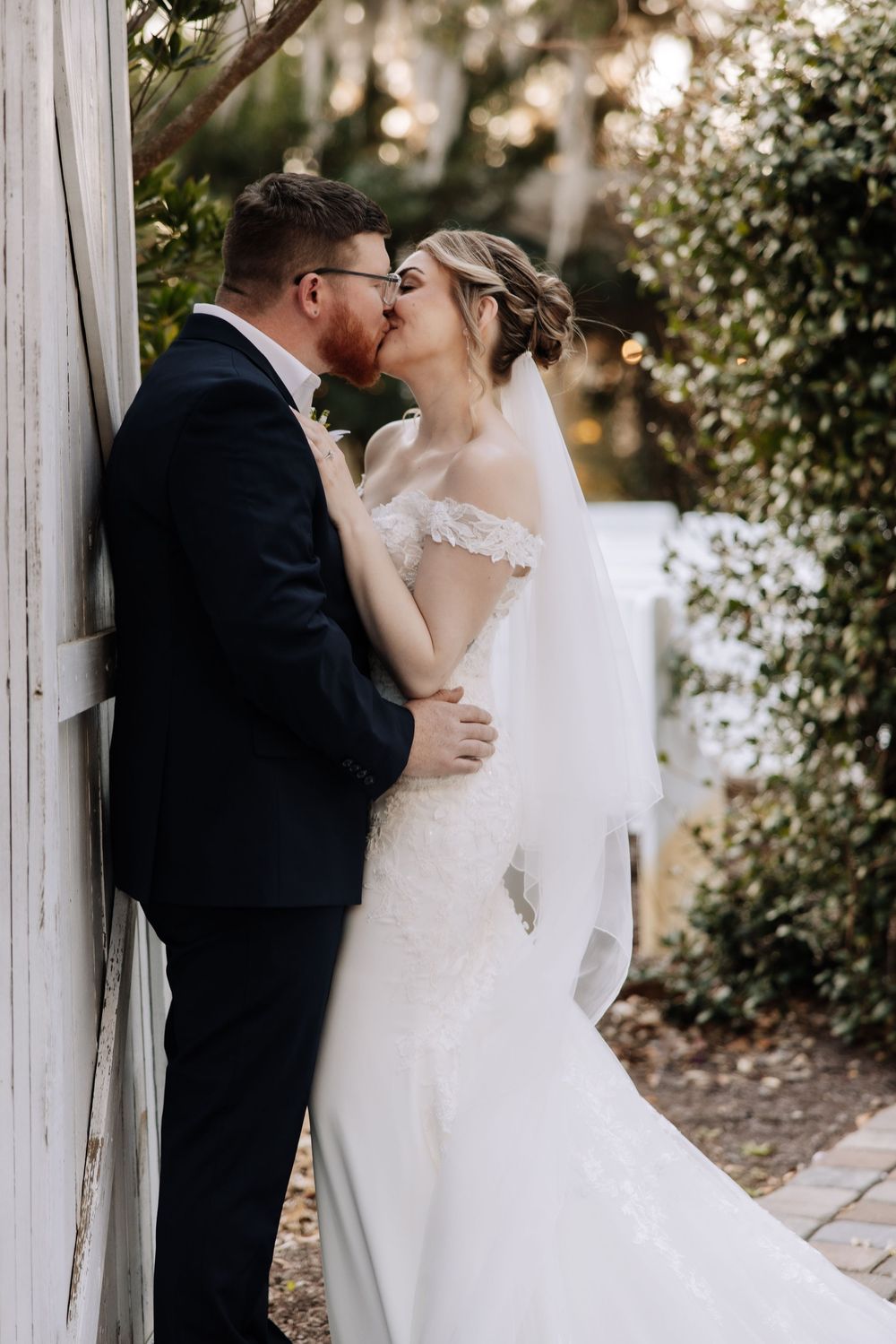 A bride and groom share a romantic kiss against a white wooden wall surrounded by lush greenery on their wedding day.