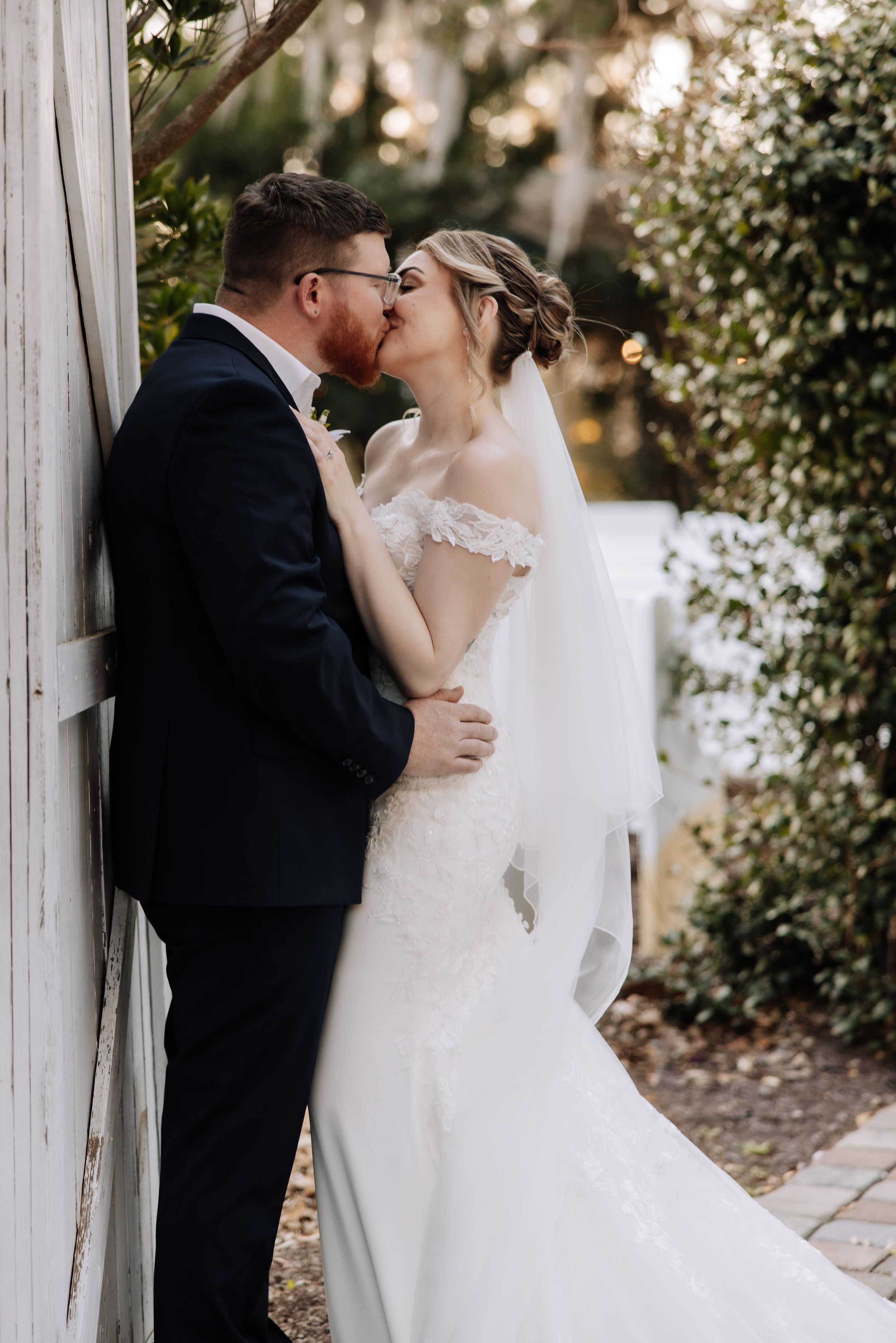 A bride and groom share a romantic kiss against a white wooden wall surrounded by lush greenery on their wedding day.