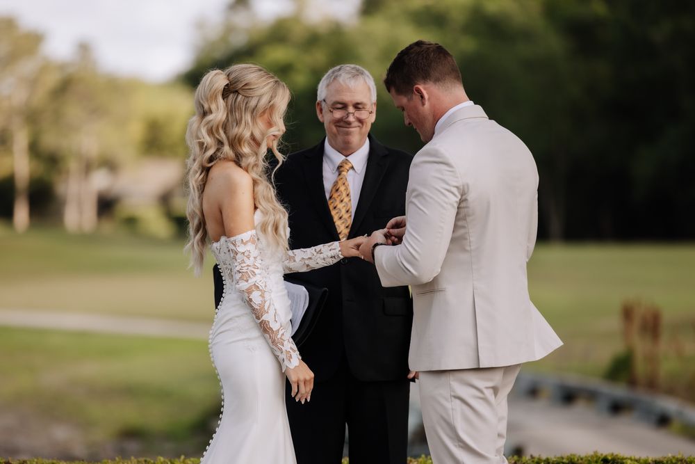 Groom placing wedding ring on bride's finger during outdoor ceremony, officiant in yellow tie standing between couple.