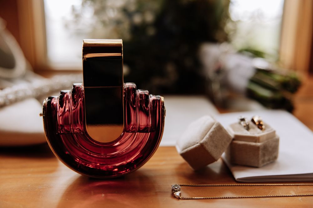 A luxurious red glass perfume bottle sits on a wooden surface beside elegant jewelry accessories.