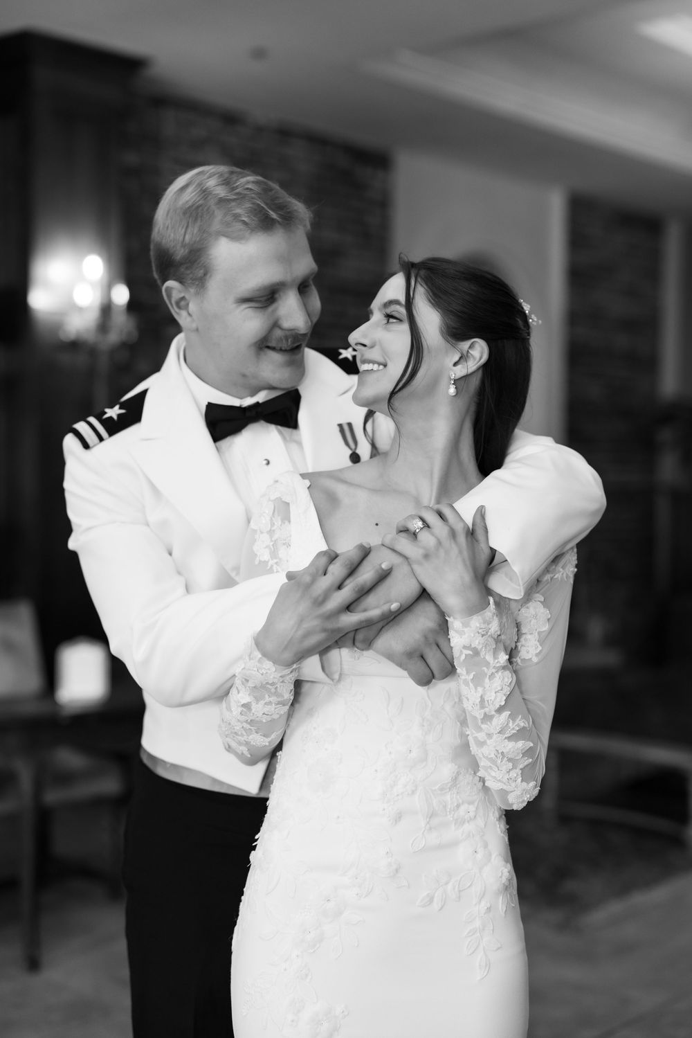 Black and white photo of a military groom in white uniform embracing his smiling bride in a lace wedding dress.