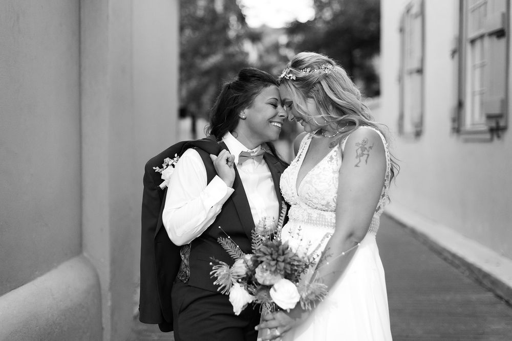 Four black and white romantic photos show tender moments between couples in wedding attire embracing near building walls.