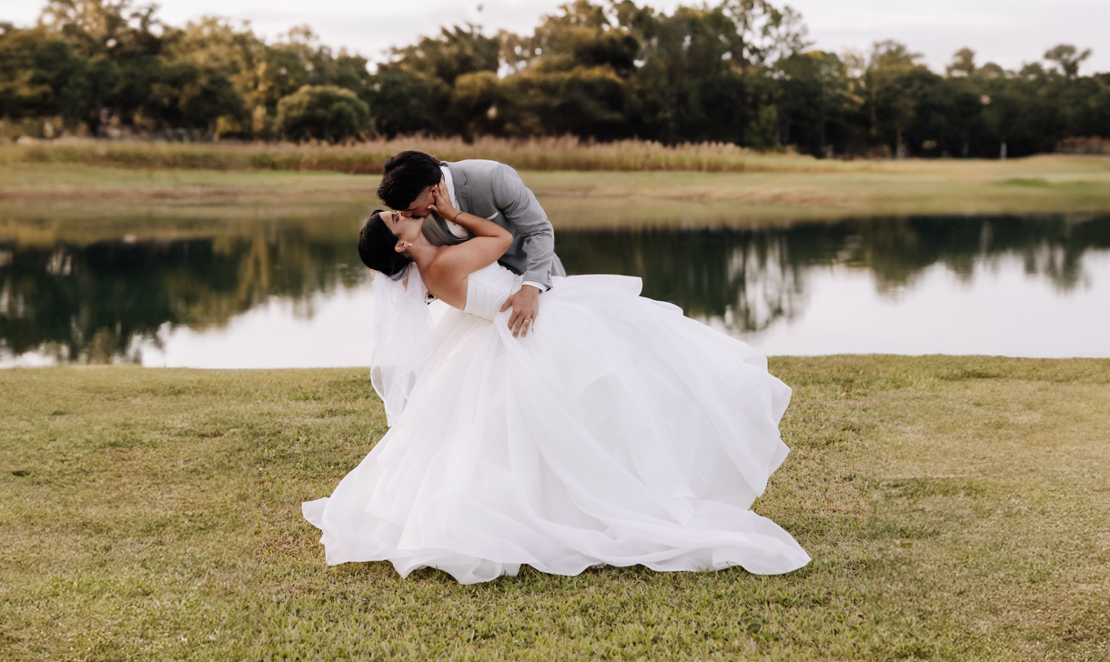 Couple shares intimate moment during lakeside wedding photo session at sunset with flowing white dress in the grass.