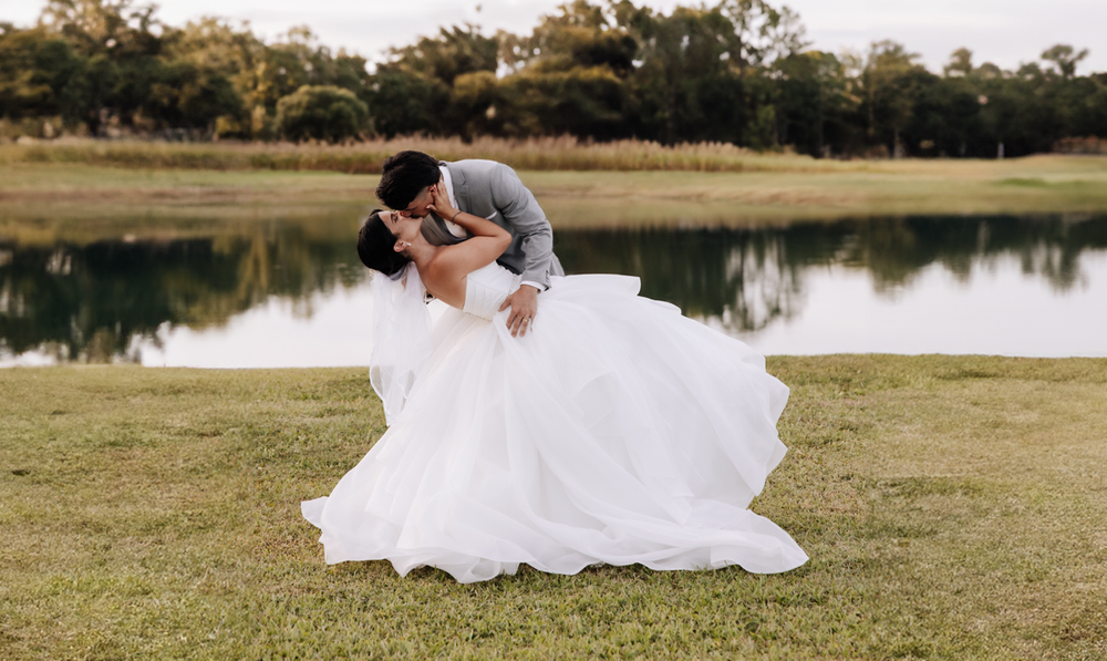 Couple shares intimate moment during lakeside wedding photo session at sunset with flowing white dress in the grass.