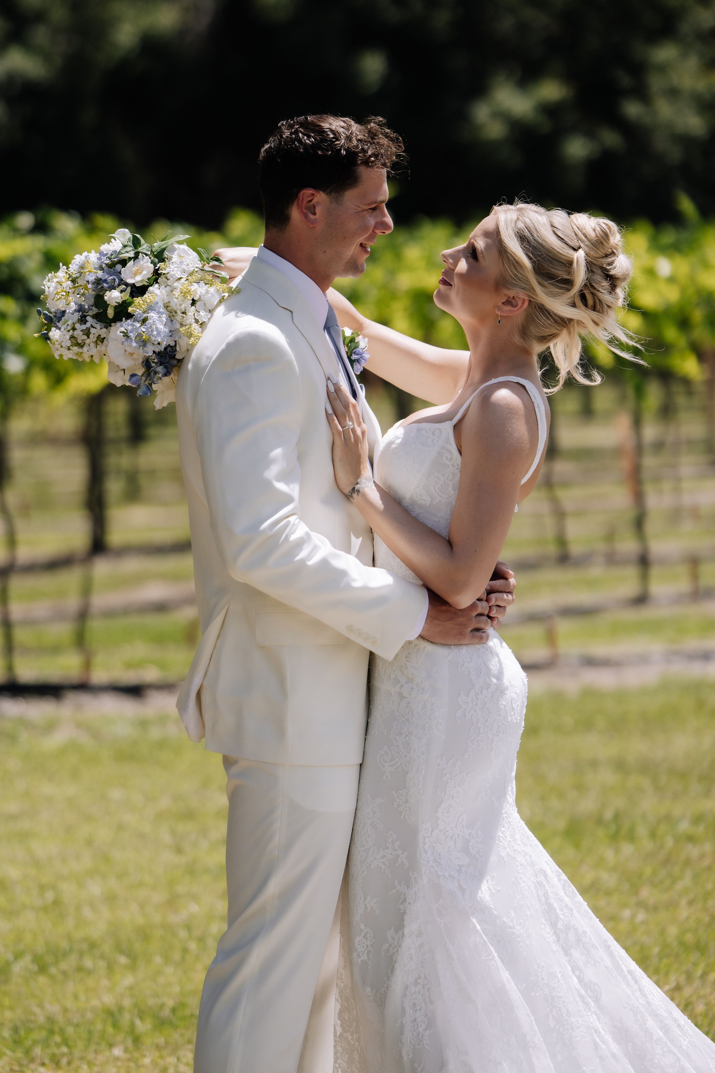Bride and groom embrace in vineyard, she holds white floral bouquet, both wearing white, smiling at each other on sunny day.