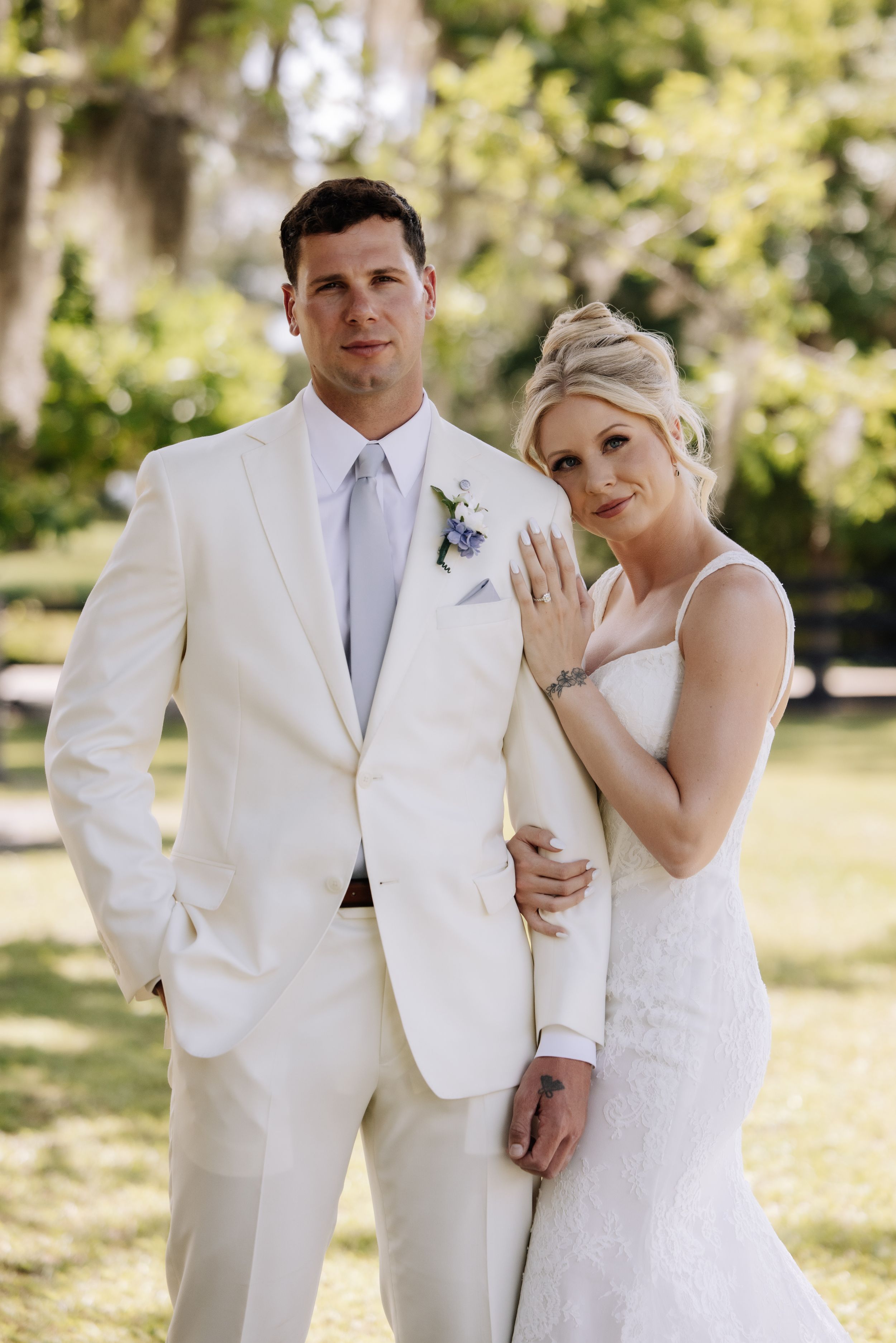 Bride and groom pose outdoors in white wedding attire, bride displaying ring, surrounded by lush green trees.