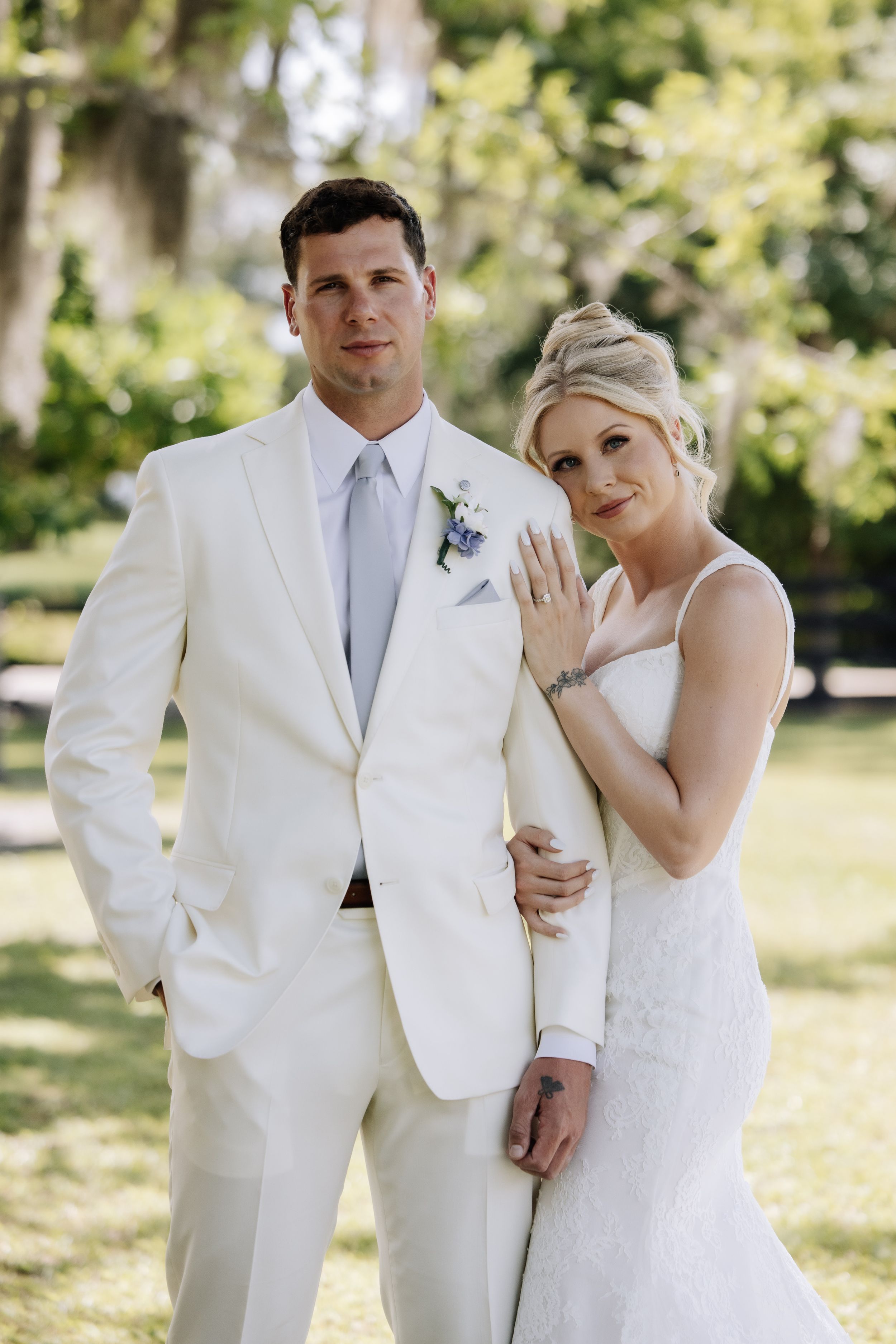 Bride and groom pose outdoors in white wedding attire, bride showing ring, surrounded by lush green trees.