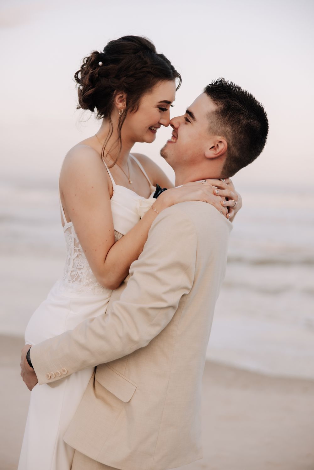 Romantic beachside moment between bride in white dress and groom in beige suit at sunset.