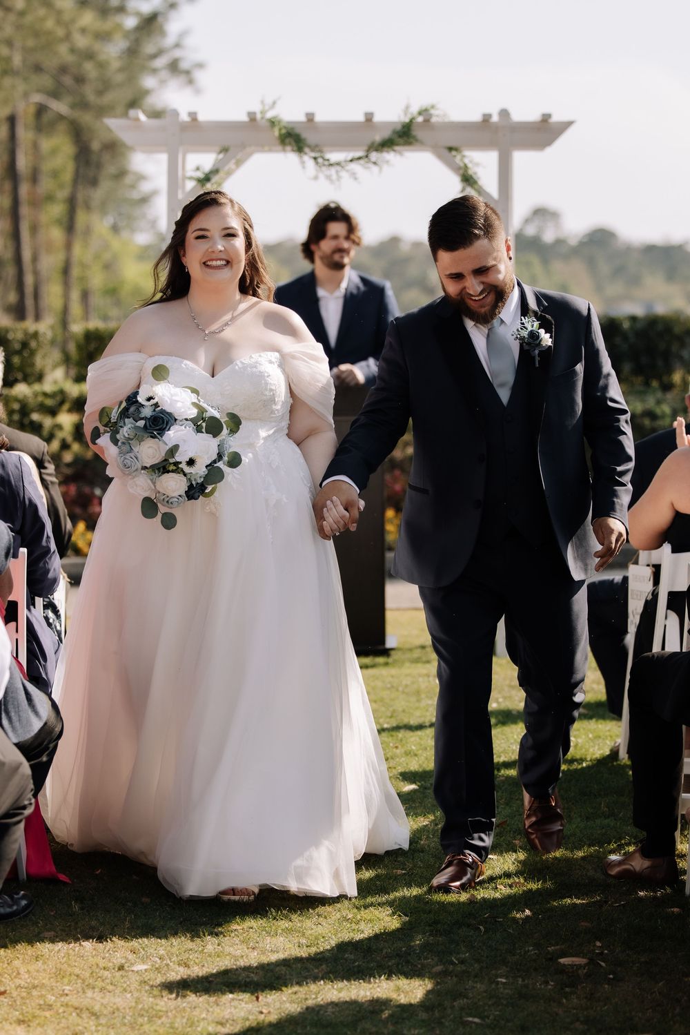 Bride and groom walk down the aisle hand-in-hand after outdoor wedding ceremony, bride holding white floral bouquet.
