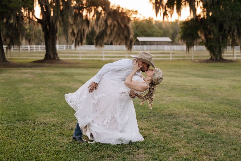 Groom in cowboy hat dips bride in white lace dress for romantic kiss on green lawn at sunset wedding.