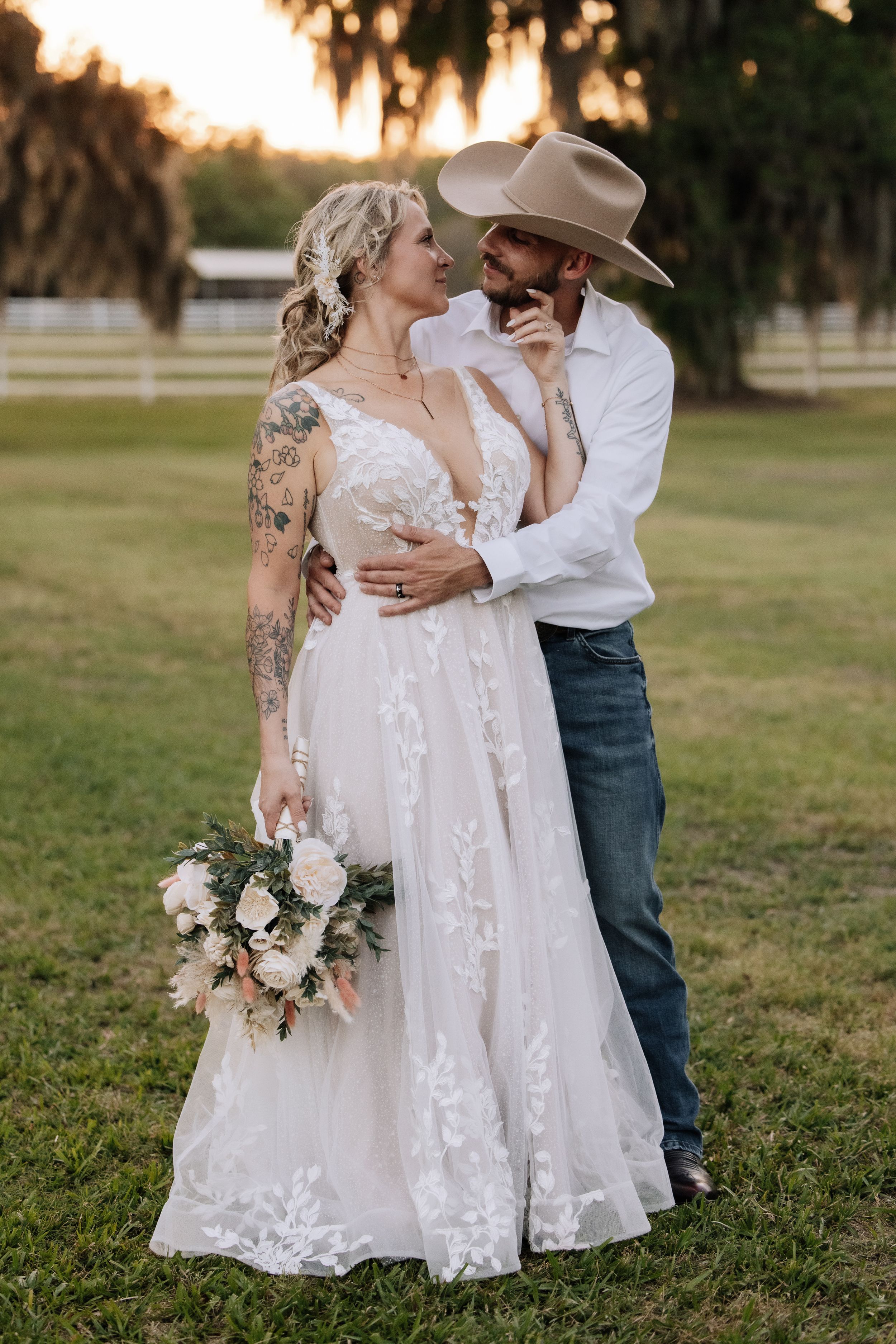A bride in lace gown holding bouquet embraces a groom in cowboy hat and jeans at a rustic outdoor wedding.