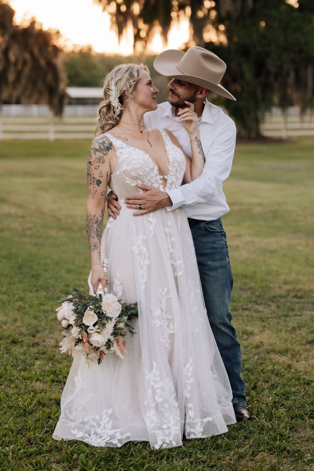 A bride in lace gown holding bouquet embraces a groom in cowboy hat and jeans at a rustic outdoor wedding.