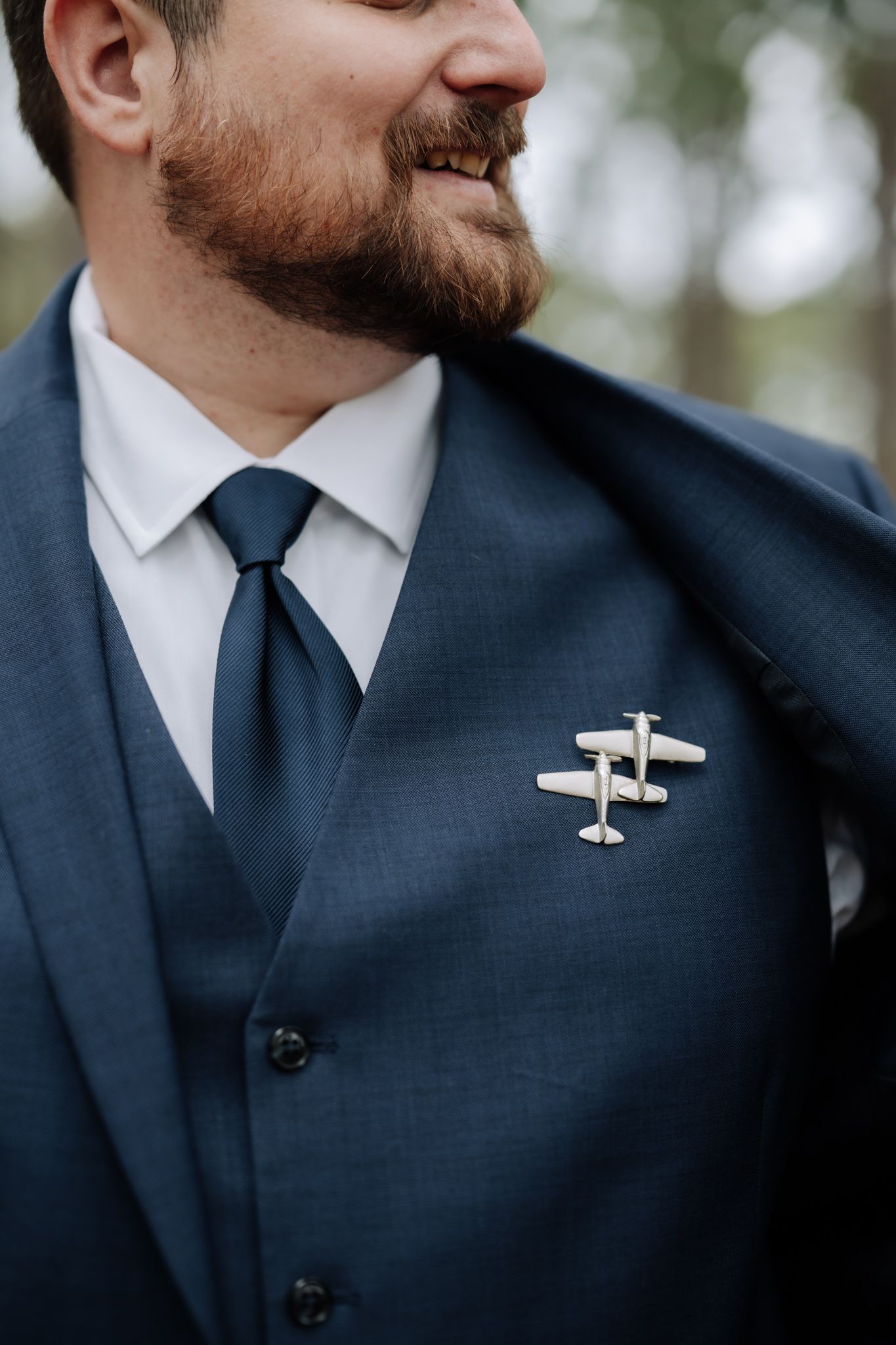 Smiling groom in navy blue suit and tie wearing a silver airplane lapel pin boutonniere at his wedding.