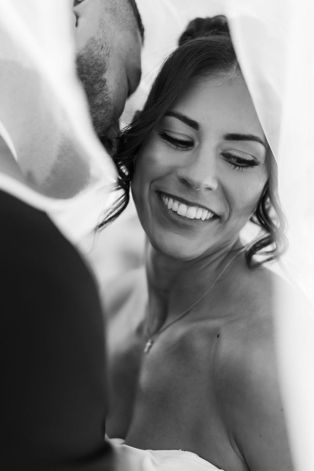 Black and white photo of a smiling bride laughing joyfully under a veil while her groom kisses her cheek.