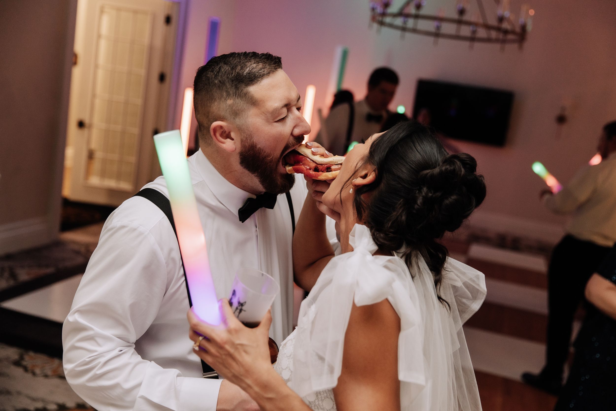 Bride and groom sharing wedding cake at reception, holding colorful glow sticks while dancing together.