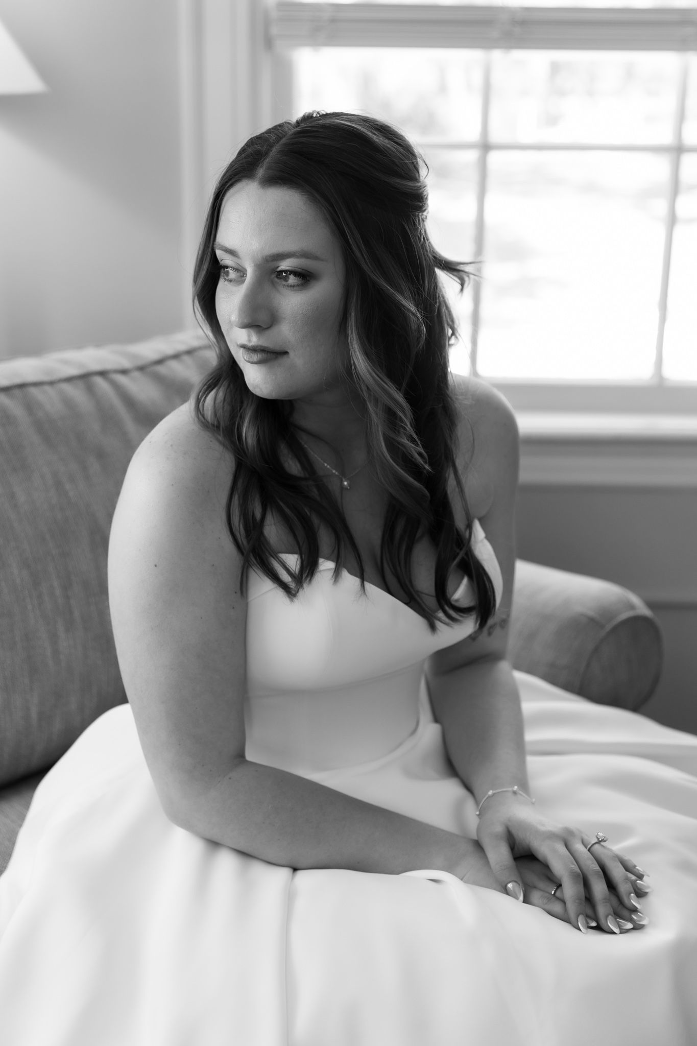 Black and white portrait of a bride in a strapless gown sitting gracefully on a sofa near a window.