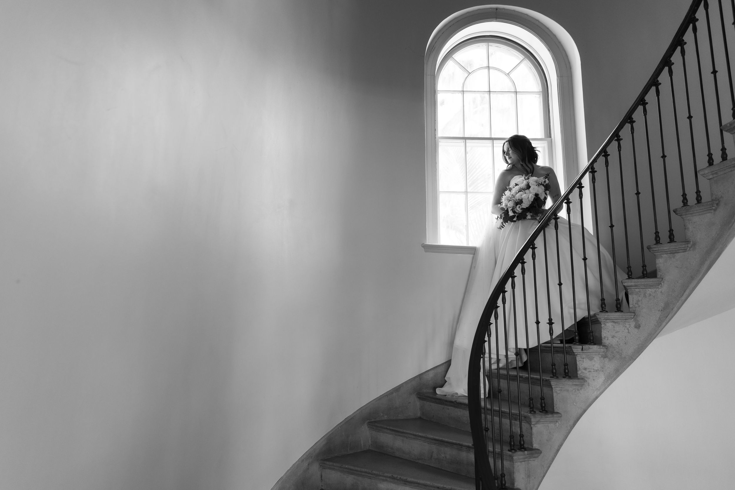 Bride in wedding gown holding bouquet stands on elegant spiral staircase by arched window, black and white photo.