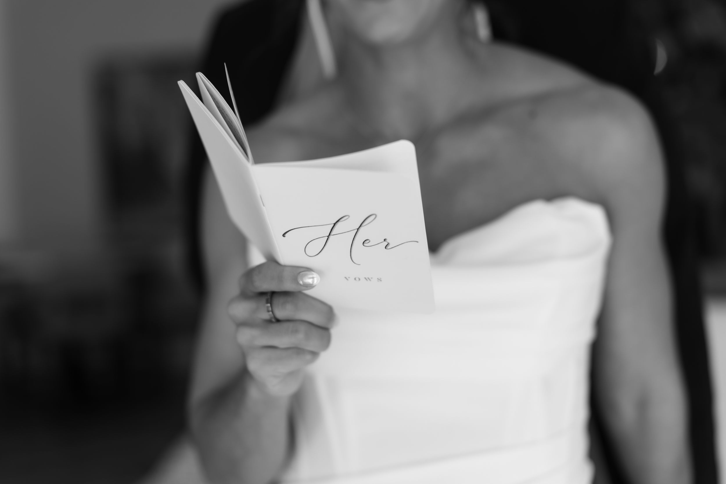 Black and white photo of a bride in a strapless gown reading wedding vows from a booklet labeled Her.