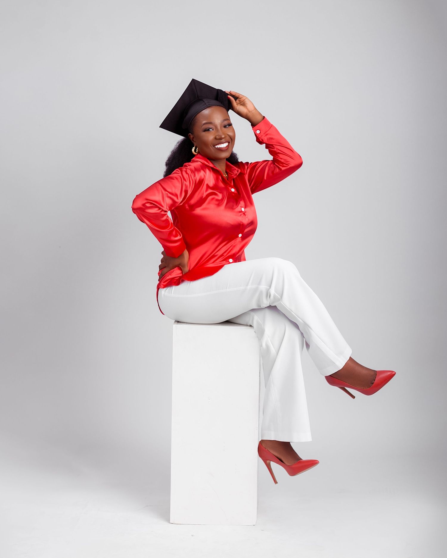 A graduate in red satin and white pants poses confidently on a white cube.