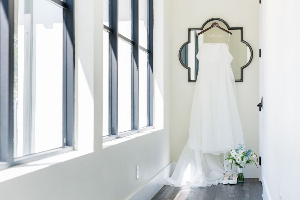 A white wedding dress hangs on an ornate wall hook near tall windows in a bright room with bouquet on floor.