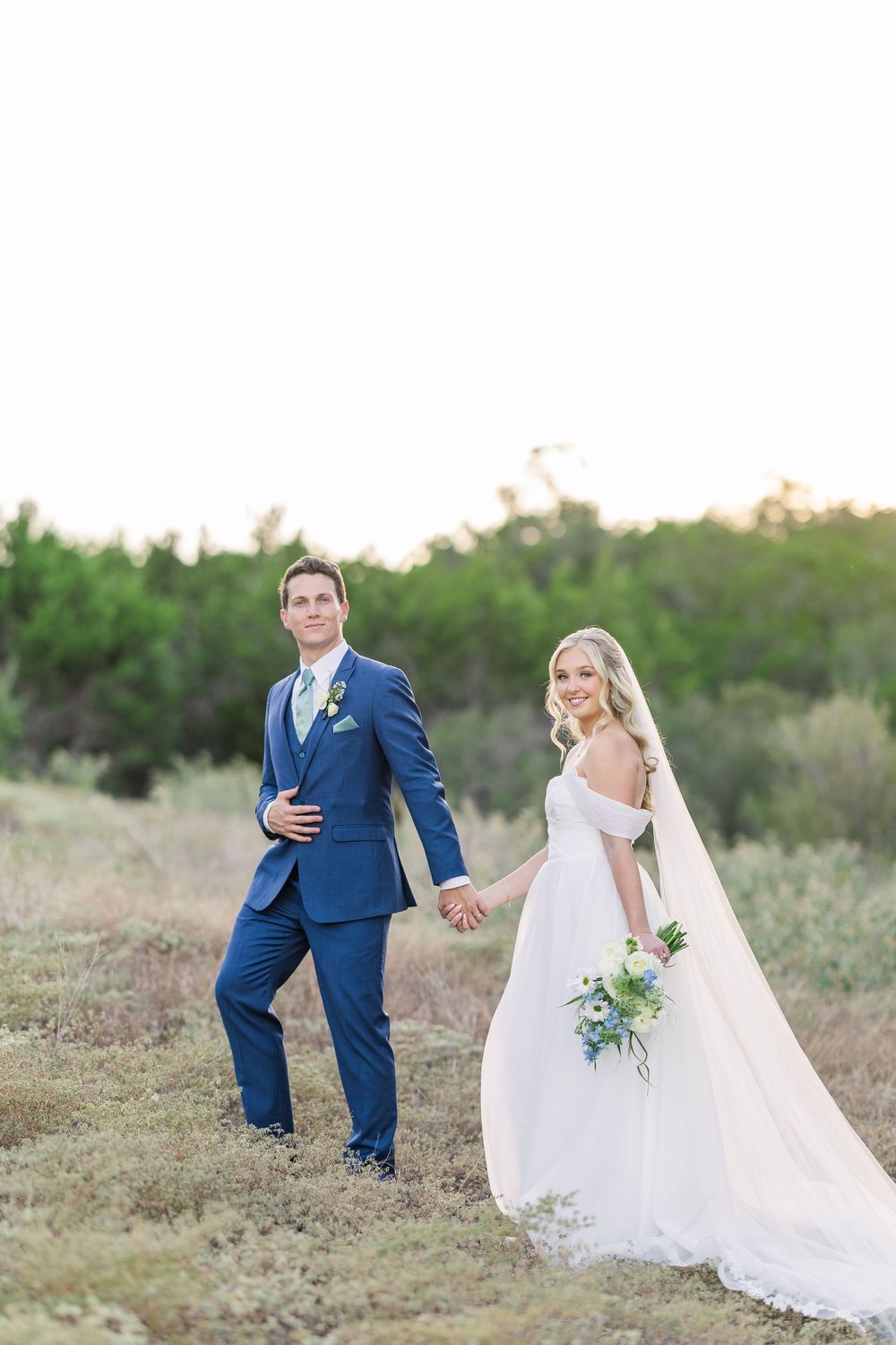 A couple in wedding attire hold hands while walking through a scenic outdoor field at sunset.