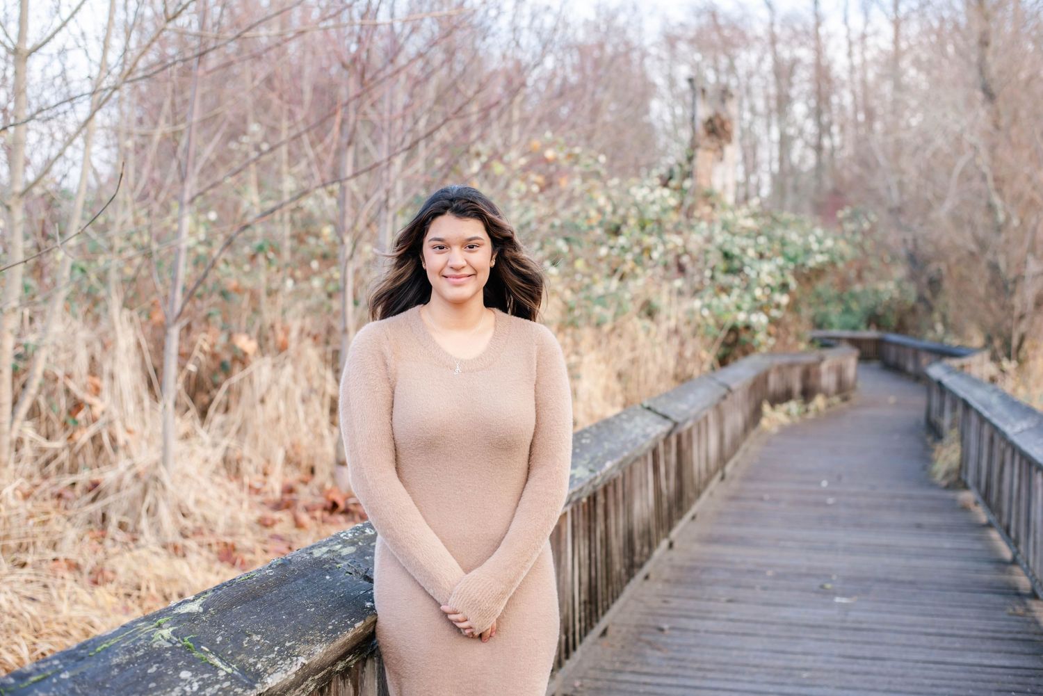 Person in neutral tones walking along a wooden boardwalk surrounded by bare winter trees.