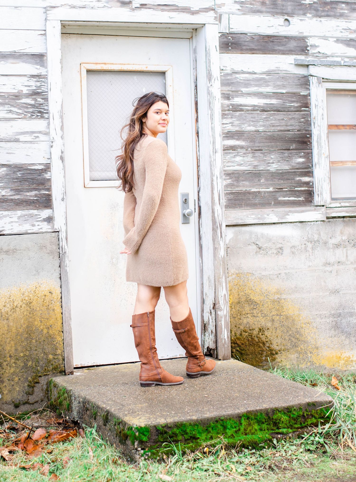 Woman in beige dress and boots standing at weathered white door of rustic building.