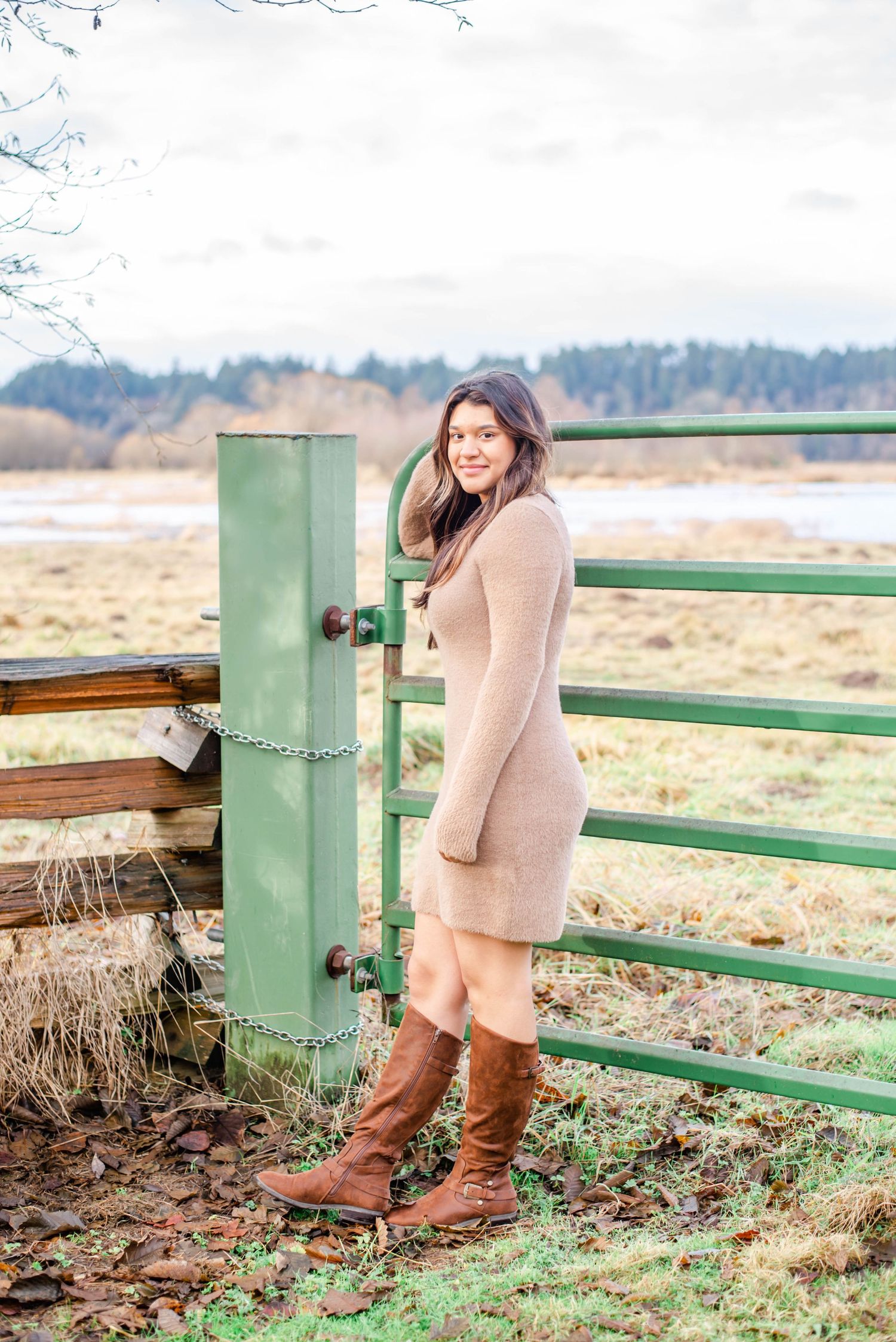 Woman in beige sweater dress standing by green metal gate post in rural landscape.