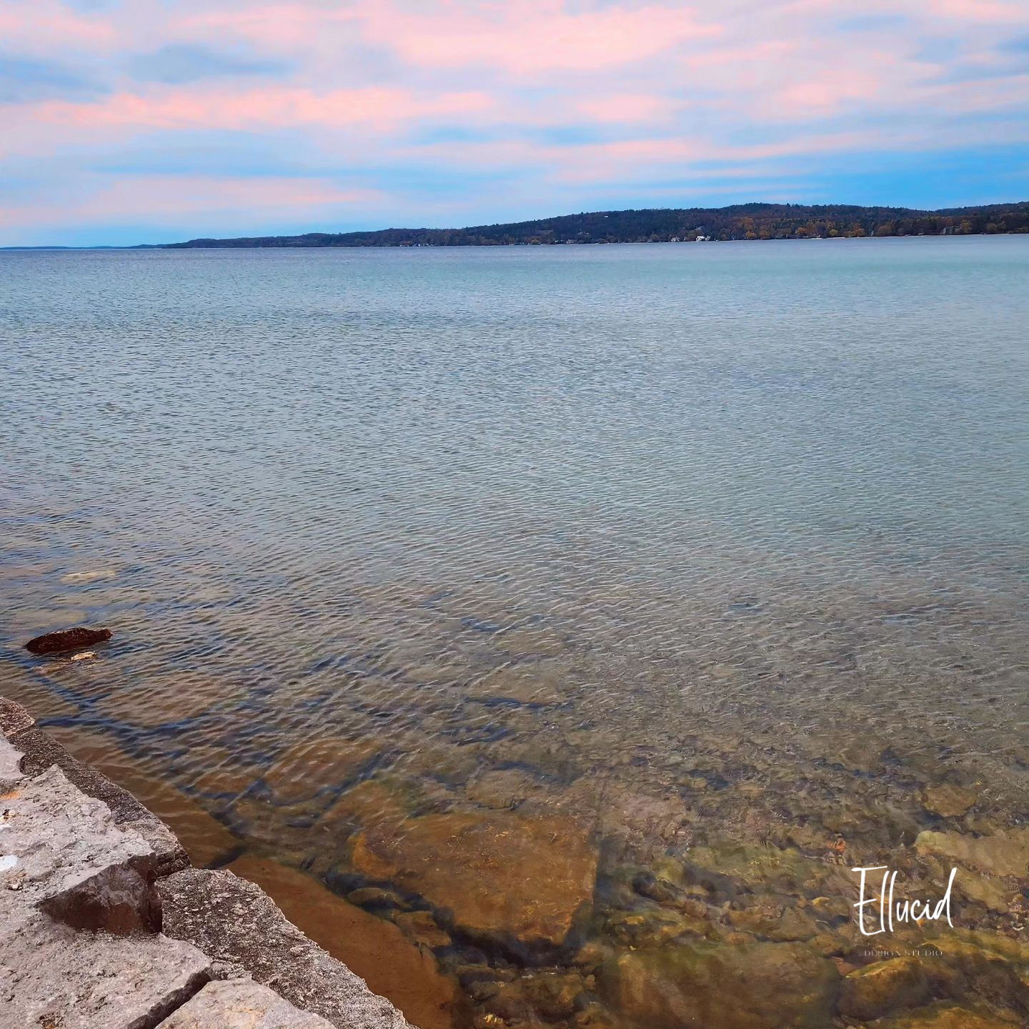 Tranquil lake view at sunset with rocky shoreline and pink-tinged clouds reflecting on calm waters.