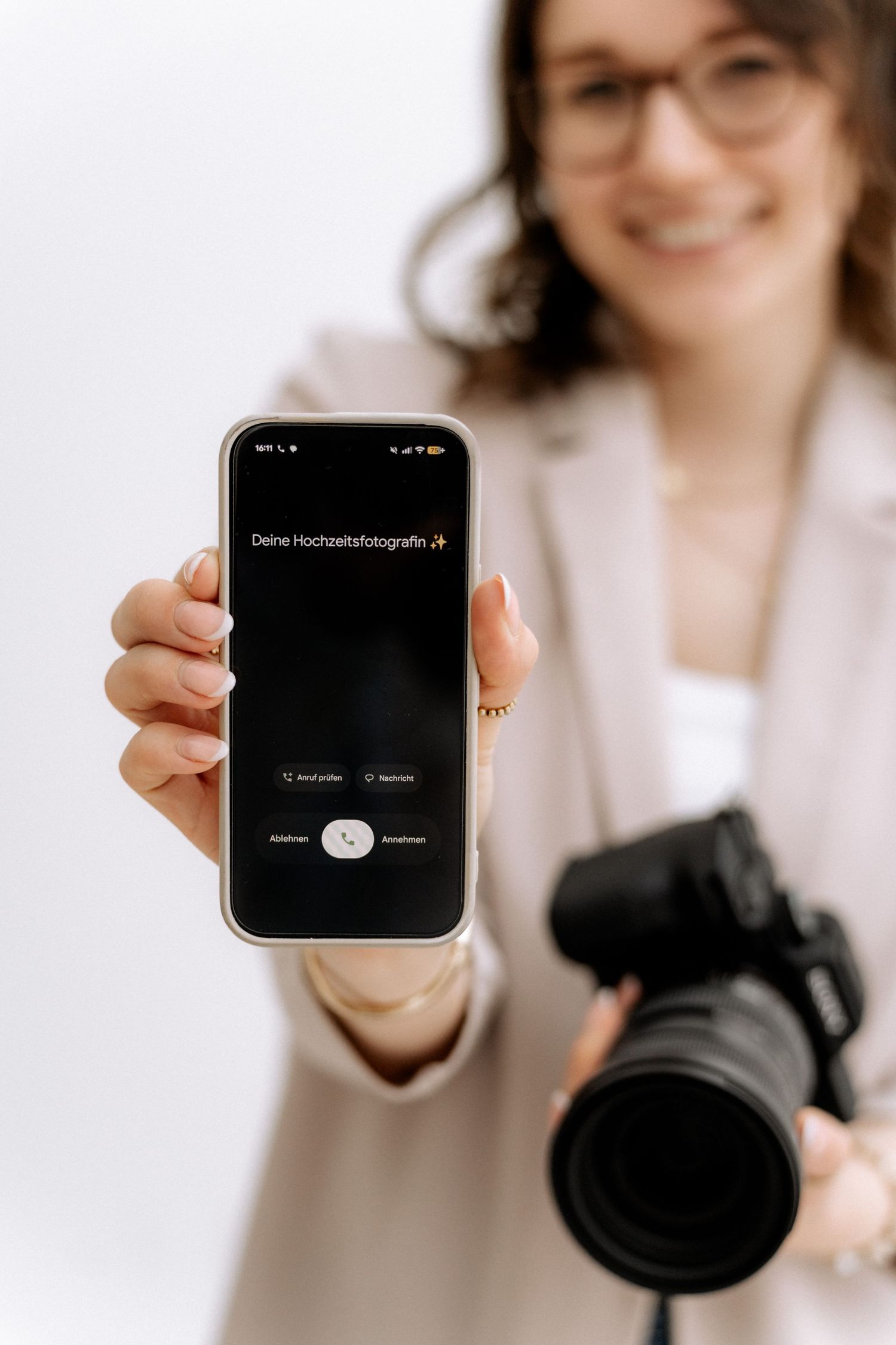 Professional photographer showing smartphone screen while holding DSLR camera with telephoto lens in white studio setting.