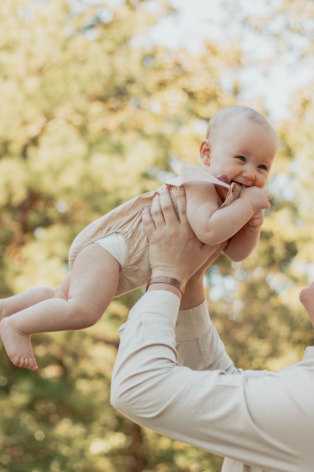 Joyful baby in beige romper being lifted in air against autumn trees.