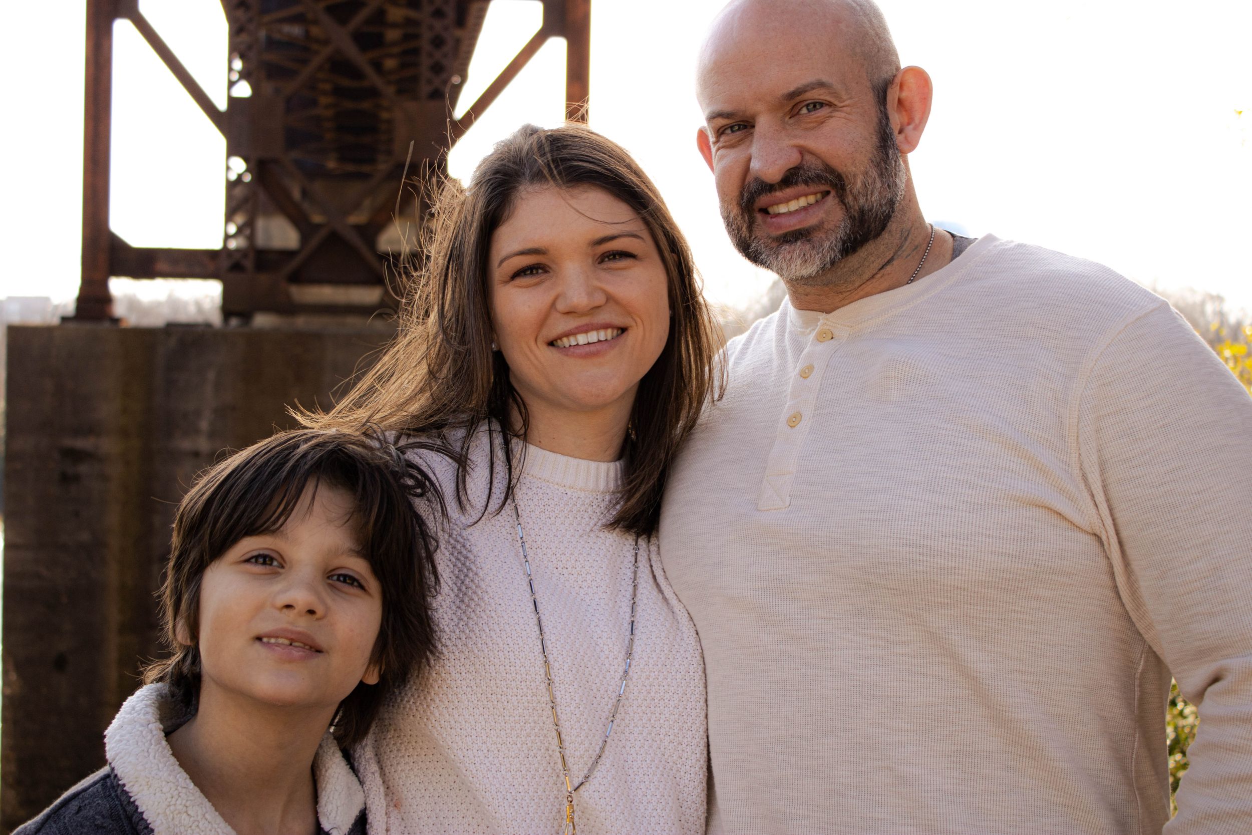 Family of three posing together outdoors near industrial structure.
